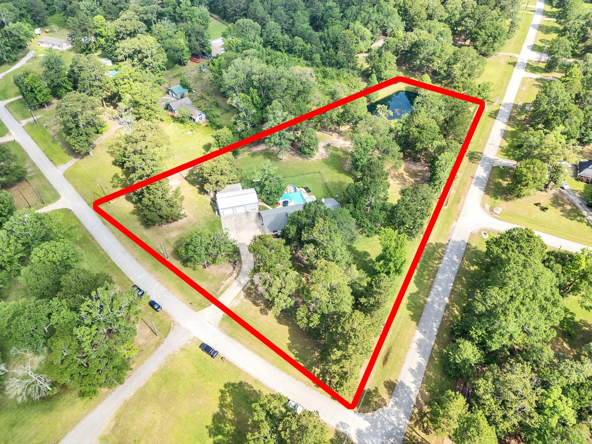 9644 Fosters Bend, Cleveland, TX 77328 This aerial photo shows a large, property outlined in red, featuring a house, pool, ample greenery, and a pond. It's situated on a corner lot with surrounding roads and mature trees, offering a serene, spacious setting. Large workshop with covered patio,concrete double drive, stunning pool, Kids play area and beautiful pond.