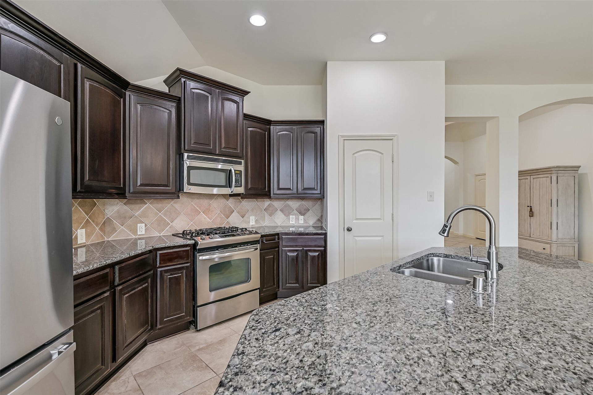 Beautiful granite countertops provide generous prep space throughout the kitchen, complemented by rich wood cabinetry & stainless steel appliances. The oversized island features a double-basin undermount sink with a high-arc faucet, while the gas range & decorative tile backsplash complete the cooking area.