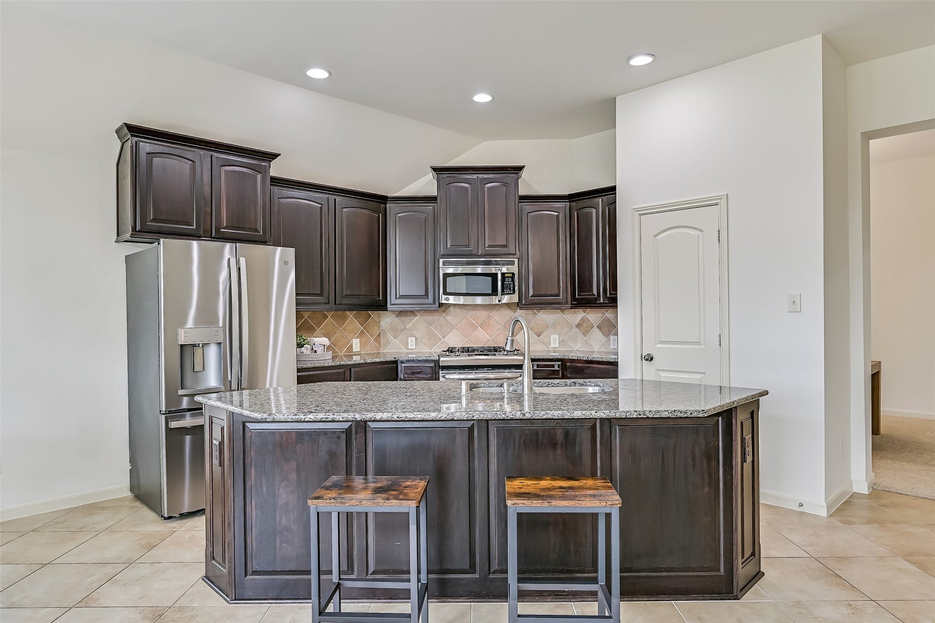 The spacious kitchen features an oversized granite island with breakfast bar seating, rich dark-stained cabinetry, stainless steel appliances, a gas range & decorative tile backsplash. Recessed lighting brightens the space, while the open design provides a seamless connection to the family room & breakfast area.