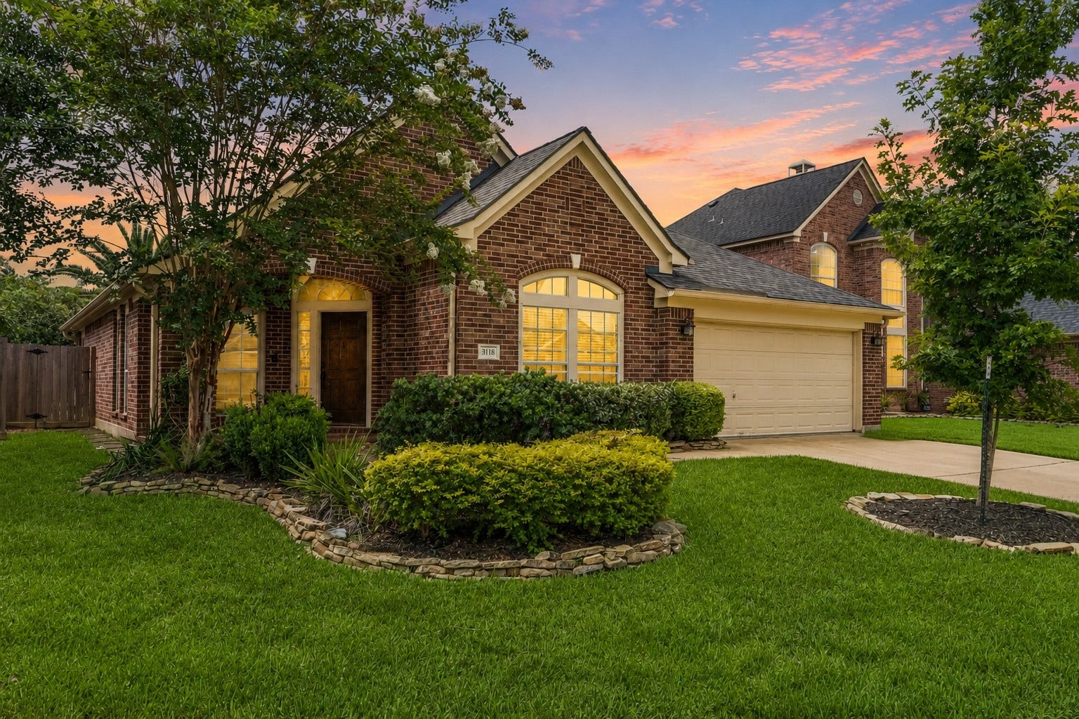 3118 Valley Court, Manvel, TX 77578 Charming brick home with a well-maintained lawn, mature trees, and attractive landscaping. Features include a two-car garage and inviting windows, all set against a picturesque sunset sky.