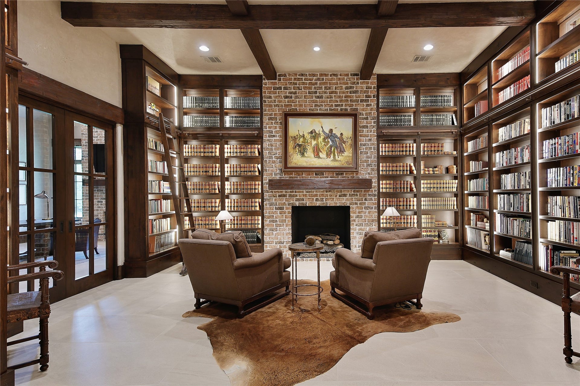 Floor to ceiling shelving, a library ladder, brick gas log fireplace, exposed beams and stone tile flooring.
