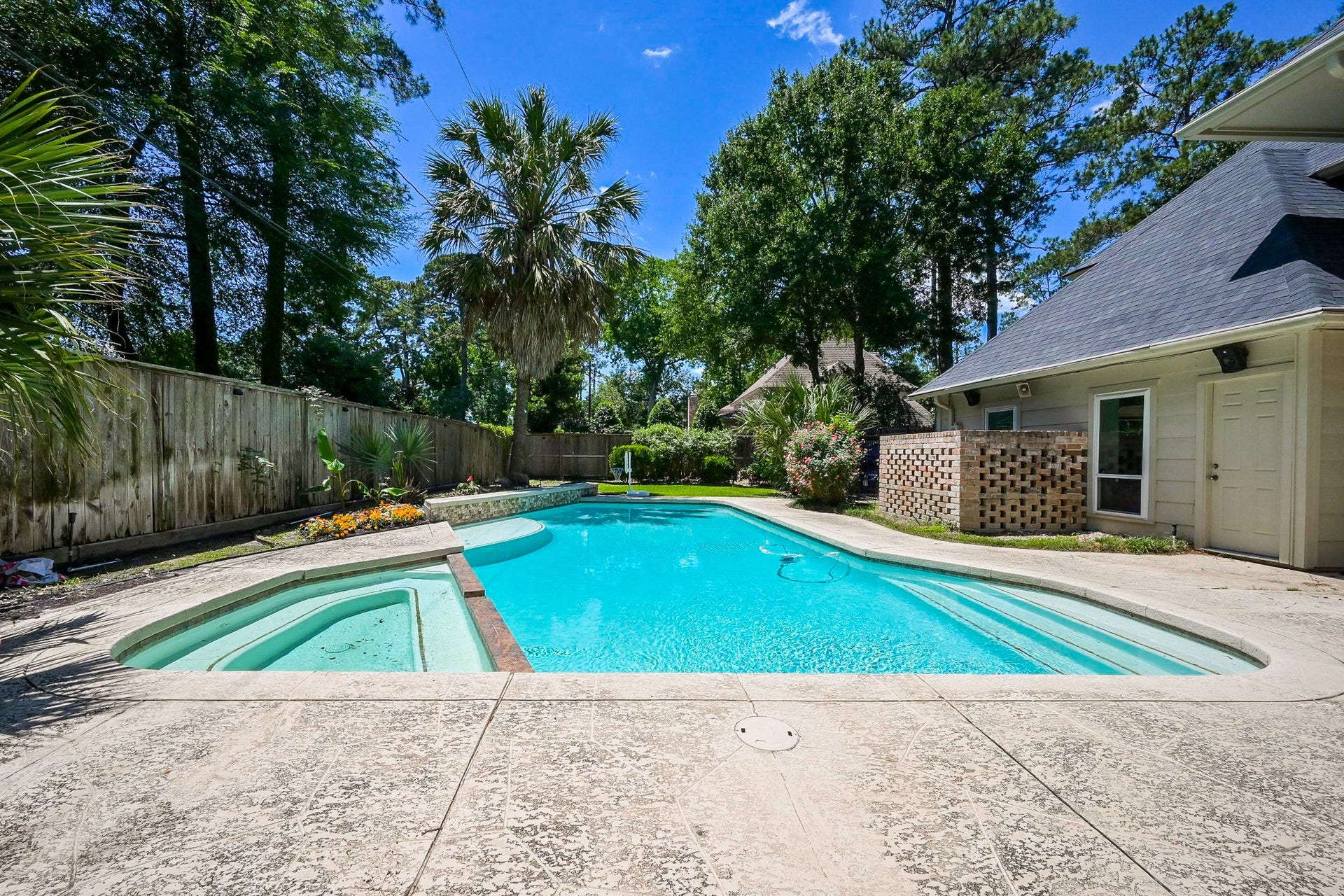 This backyard features a spacious pool with a built-in hot tub, surrounded by a concrete patio. Lush greenery and tall trees provide privacy, and a wooden fence encloses the area.