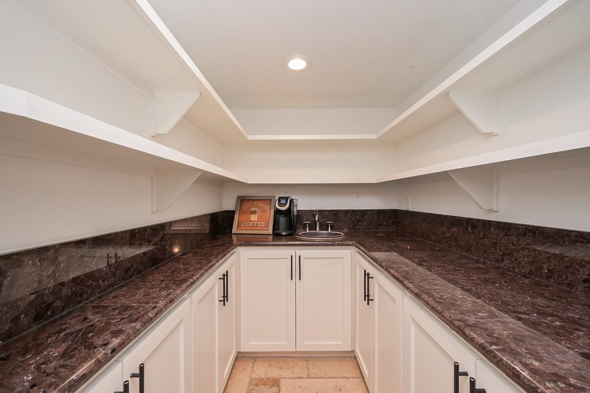 A spacious wet bar/pantry with elegant dark granite countertops, ample white cabinetry, and open shelving. Ideal for storage and organization, featuring a small sink and a coffee maker area.