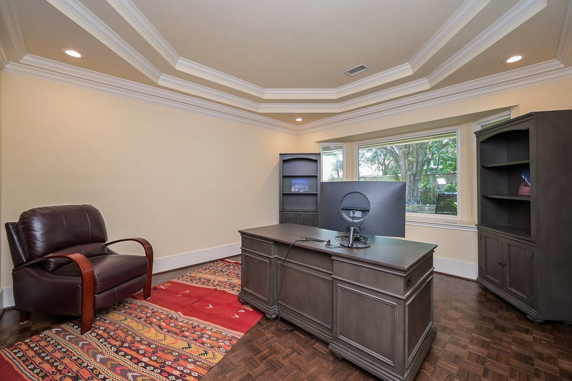 This formal den could also be used as a spacious home office. It has a coffered ceiling with recessed lighting and a large window providing natural light and a view of the greenery outside.