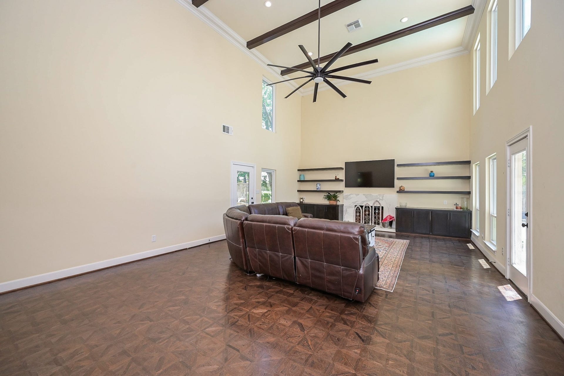 This spacious living room features high ceilings with exposed beams, large windows for natural light looking out to the pool. It includes a fireplace with built-in shelves creating a perfect space for relaxation and entertainment.