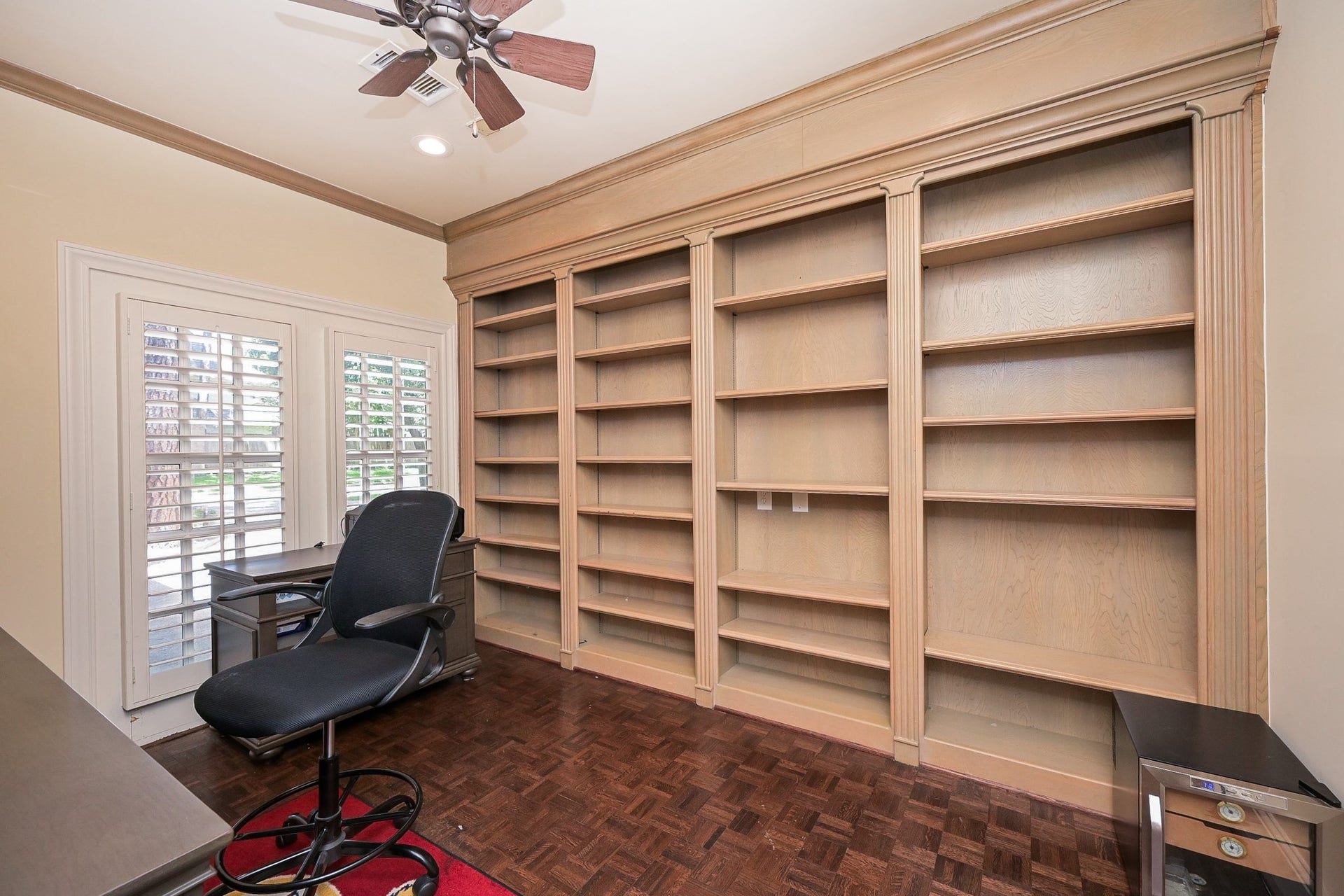 This photo showcases a home office with built-in wooden bookshelves. The room is well-lit with natural light from large windows with shutters. The parquet flooring and ceiling fan add a touch of elegance.
