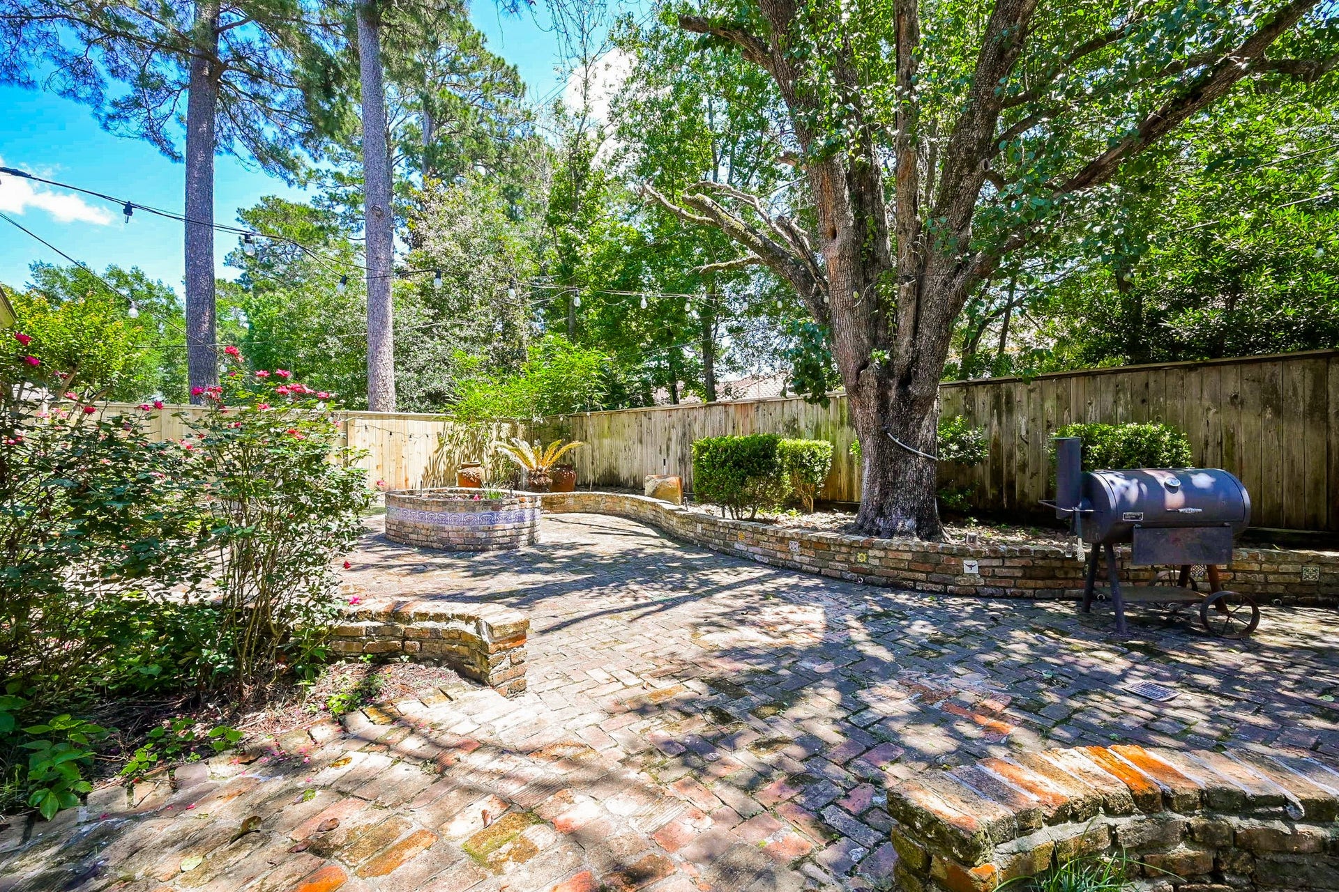 This charming backyard features a spacious brick patio with a built-in fire pit, mature trees providing shade, and a smoker for outdoor cooking. The space is enclosed by a wooden fence, offering privacy and a cozy atmosphere for relaxation or entertaining.