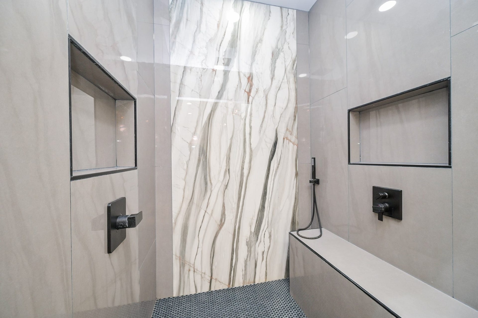 This is a sleek, modern shower with large marble-patterned tiles, built-in shelving, and a bench. The minimalist design features a handheld showerhead and matte black fixtures, creating a luxurious spa-like feel.