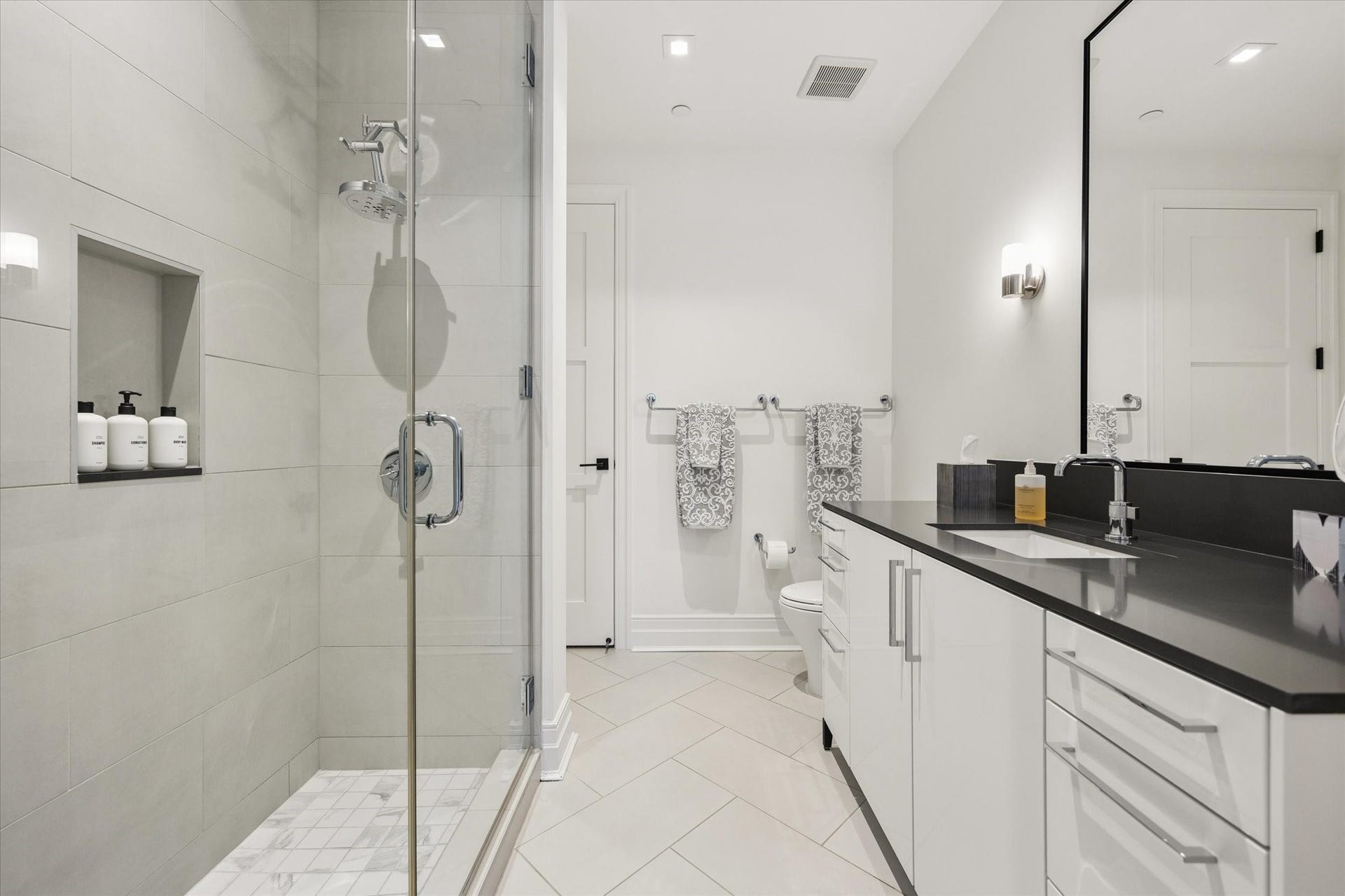 6017 Memorial Drive, #505, Houston, TX 77007 This elegantly finished secondary bathroom features a spacious walk-in shower with frameless glass enclosure, contemporary porcelain tile, and polished chrome fixtures. A sleek vanity with contrasting dark countertop, generous cabinet storage, and an oversized framed mirror creates a sophisticated yet practical setting. Thoughtful details, including a recessed shower niche and modern wall sconces, reflect the residence's commitment to quality and refined design.