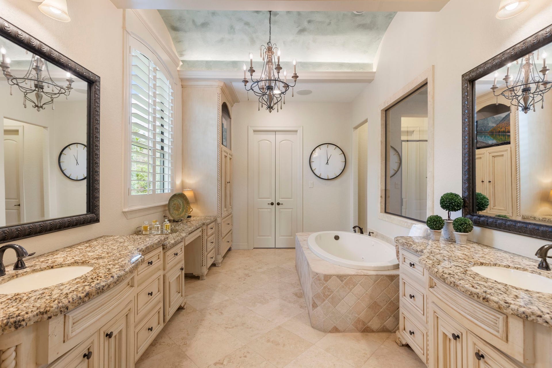 Spa-inspired Primary Bath with French door entry, featuring Travertine flooring, barrel ceiling with crown molding and accent lighting, decorative chandelier, and integrated sound system. Includes a jetted soaking tub, oversized walk-in shower with bench and tumbled stone accents, dual vanities with granite counters, seated vanity area, built-in linen storage, and an expansive walk-in closet with custom shelving.