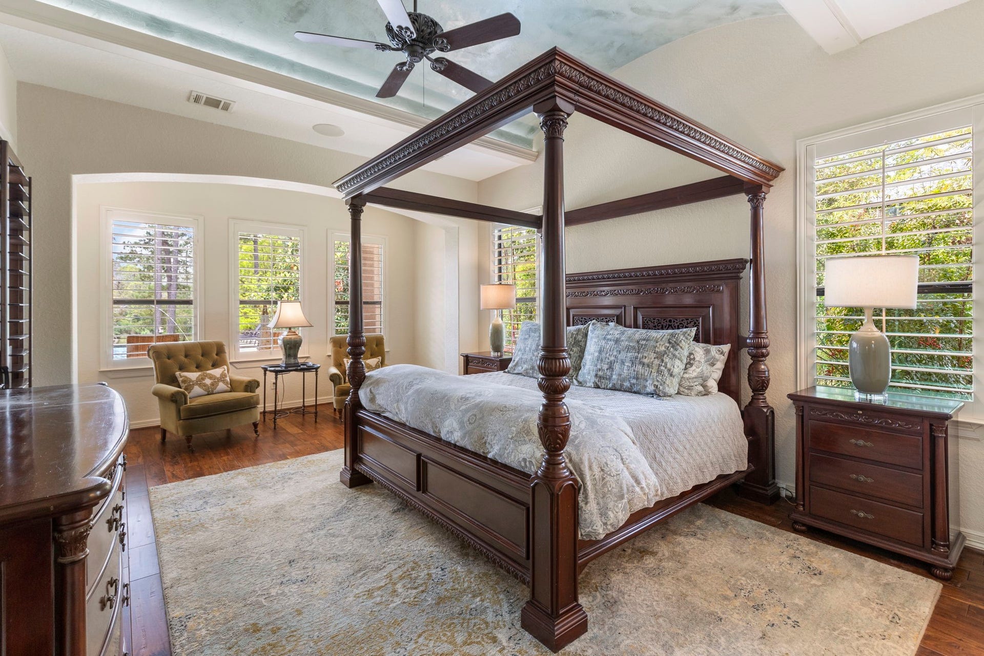 Spacious downstairs Primary Suite with rich wood flooring and an elegant barrel ceiling with crown molding and accent lighting. Features a sitting area, wall of windows overlooking the pool and lake, sound system, ceiling fan, and private patio access.