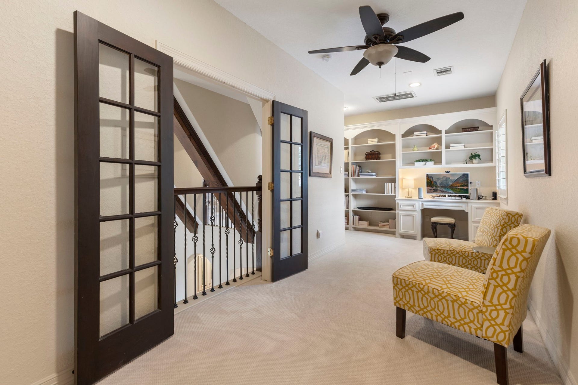 Inviting upstairs library retreat with arched custom built-ins, integrated desk, updated carpet, ceiling fan, and a charming Juliet balcony with French doors - a perfect reading or homework space.