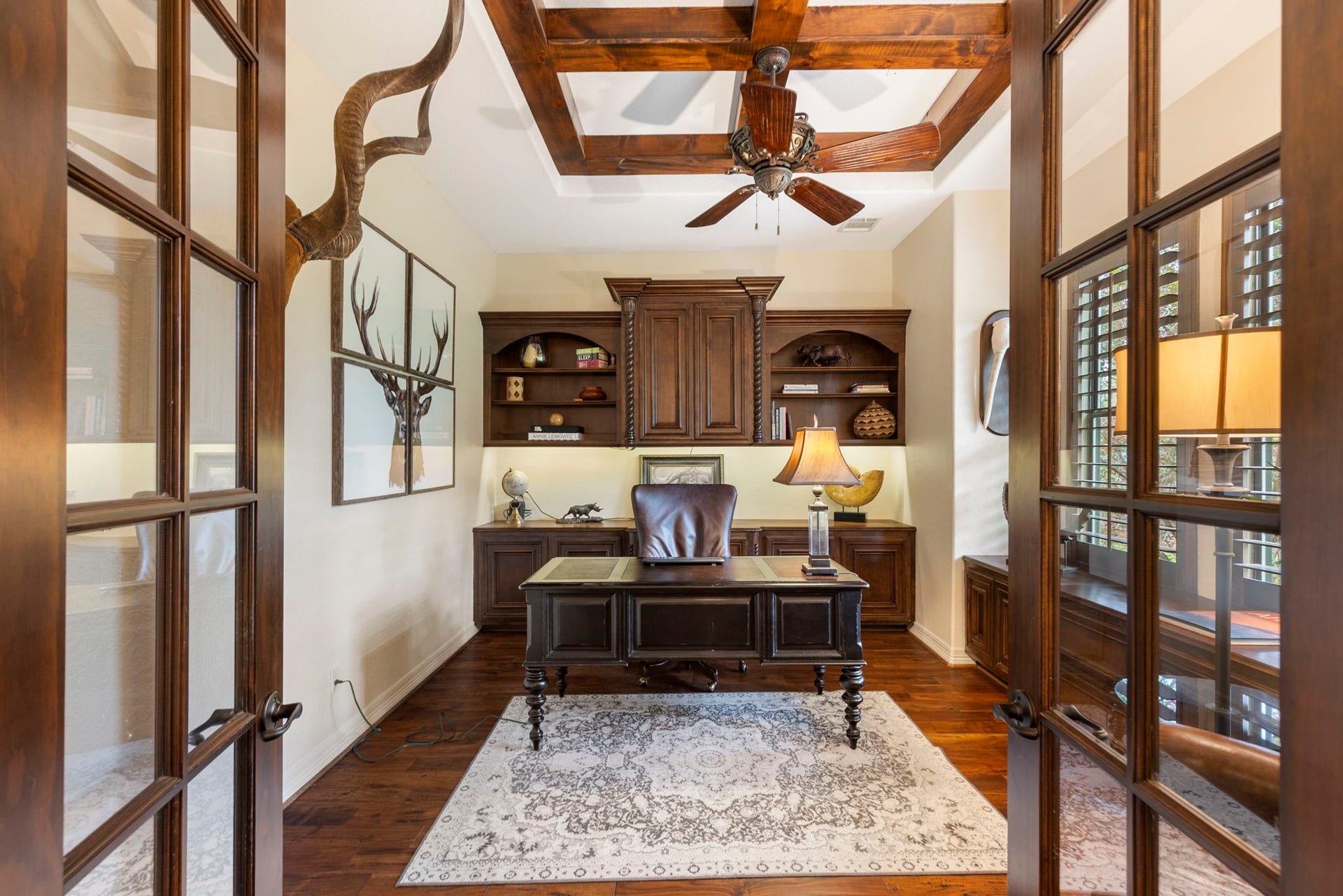 Distinguished Executive Study with glass French doors, wood flooring, and a striking beam-coffered ceiling. Custom built-ins and credenza provide refined functionality, complemented by plantation shutters and a ceiling fan.
