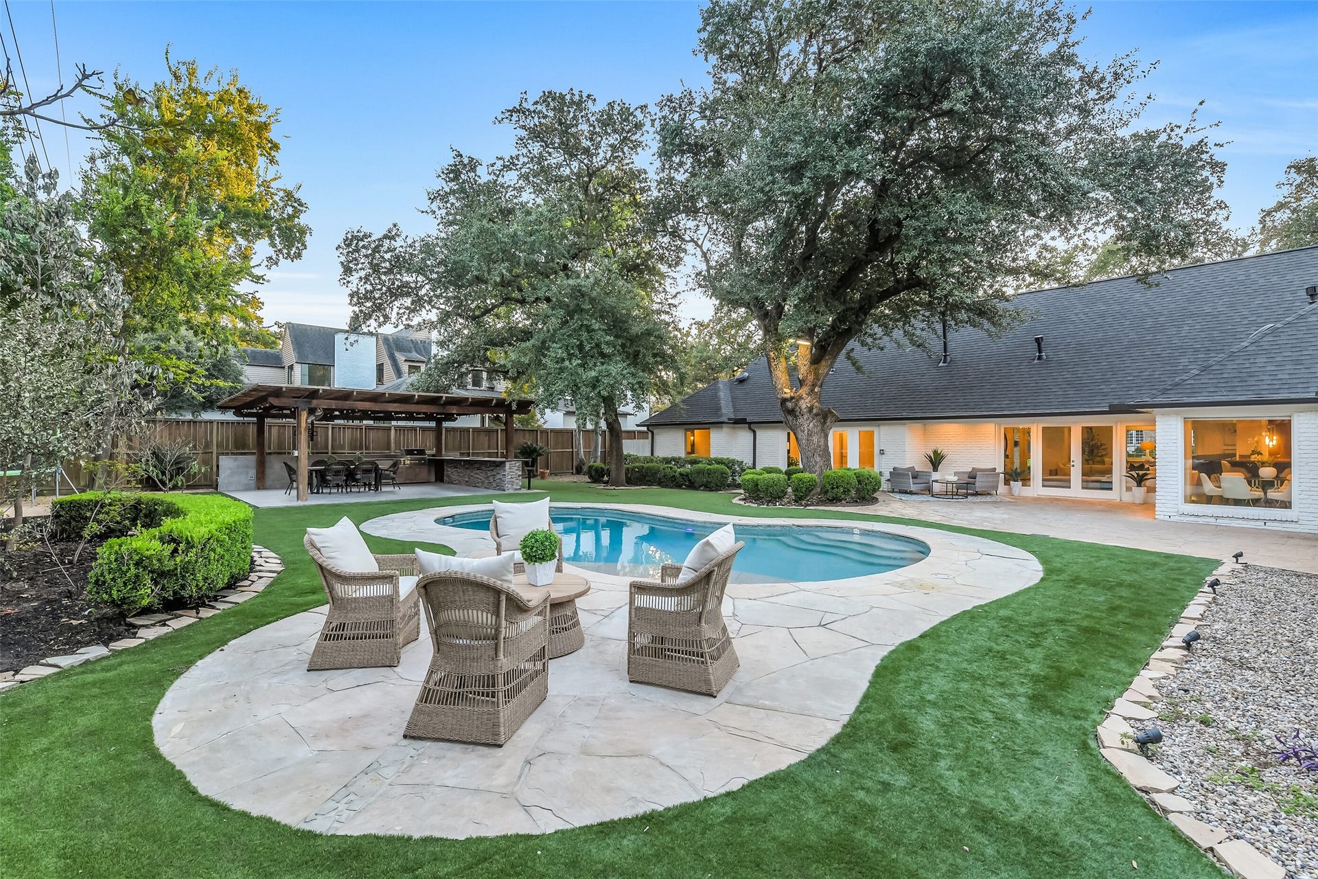 Beautifully landscaped backyard offers a private outdoor retreat with a sparkling pool, expansive stone decking, mature trees, multiple seating areas, and a covered outdoor kitchen perfect for entertaining.