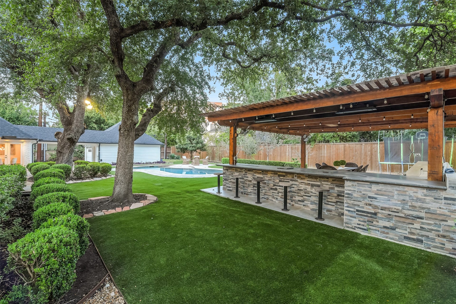 Another view of the beautifully maintained backyard showcasing lush landscaping, mature shade trees, a sparkling pool, and an expansive covered outdoor kitchen with bar seating.