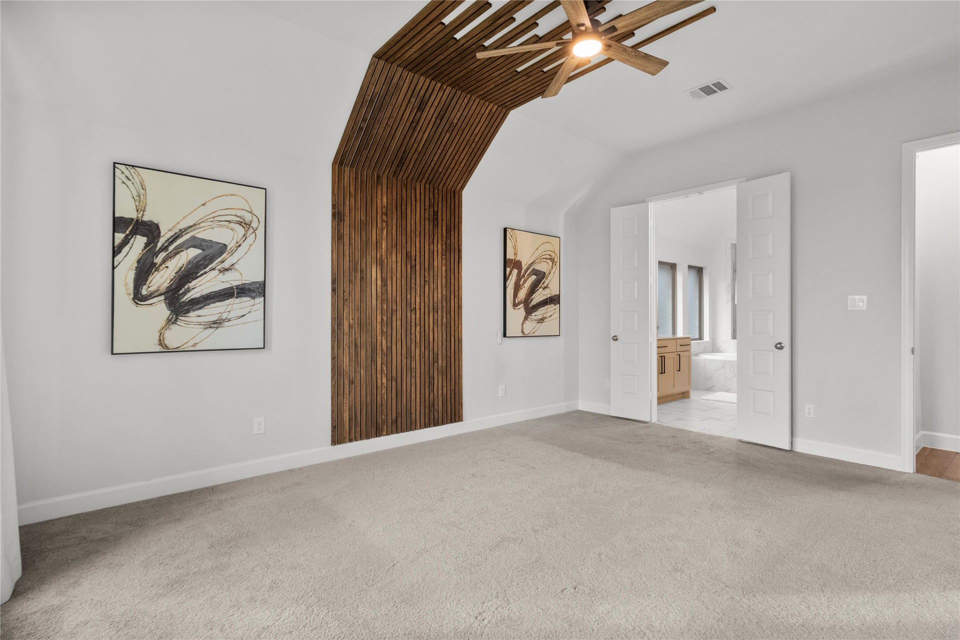 9934 Starry Night Lane, Manvel, TX 77578 Spacious bedroom with a unique wood accent wall and modern ceiling fan, featuring double doors leading to a bright en-suite bathroom.