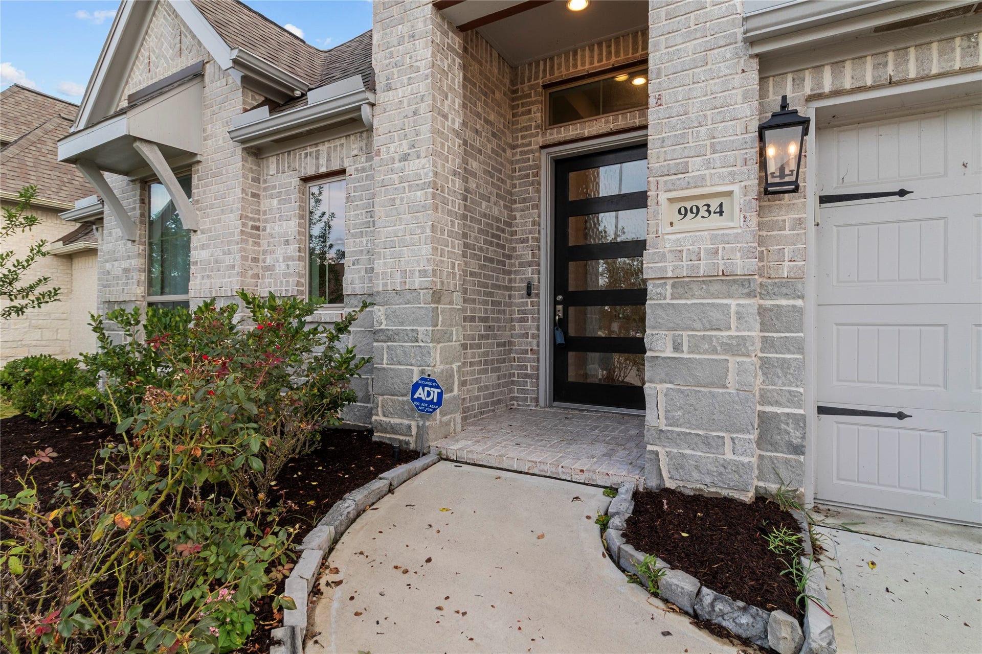 9934 Starry Night Lane, Manvel, TX 77578 Charming entrance with a modern glass door, elegant brickwork, and a welcoming porch, complemented by tasteful landscaping and a classic lantern-style light fixture.
