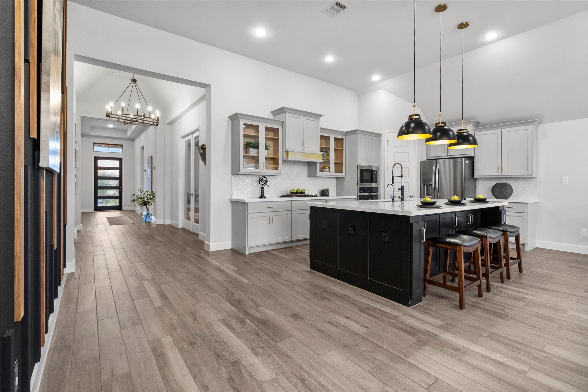 9934 Starry Night Lane, Manvel, TX 77578 Spacious modern kitchen with sleek cabinetry, a large island with seating, and elegant pendant lighting, seamlessly flowing into a welcoming hallway adorned with stylish chandeliers and abundant natural light.