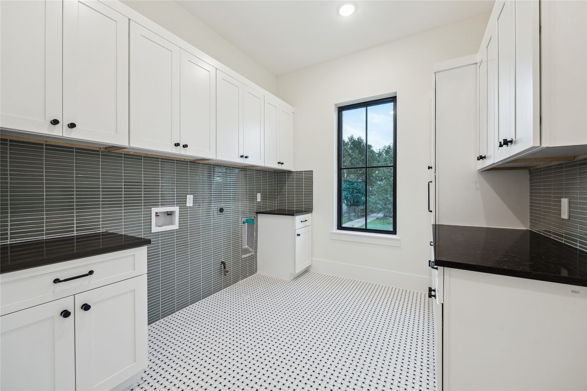 431 W 30th Street, Houston, TX 77018 This spacious upstairs laundry room features sleek black countertops, white cabinetry, and a modern gray tile backsplash. The room is well-lit with natural light from a large window, and has patterned tile flooring, offering a clean and functional space.