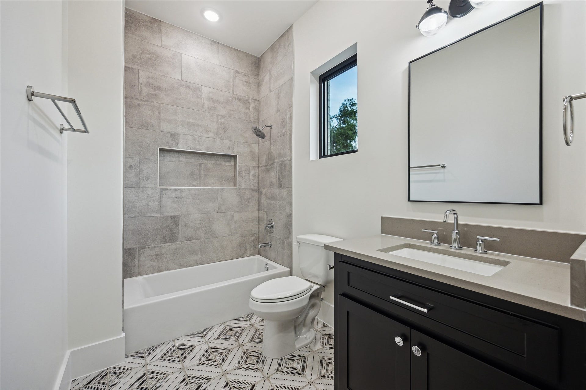431 W 30th Street, Houston, TX 77018 This modern bathroom features a sleek design with a spacious vanity, dark cabinetry, and a rectangular mirror. It includes a bathtub with gray tile surround and inset shelving. The floor has a geometric tile pattern, and there's a window for natural light.