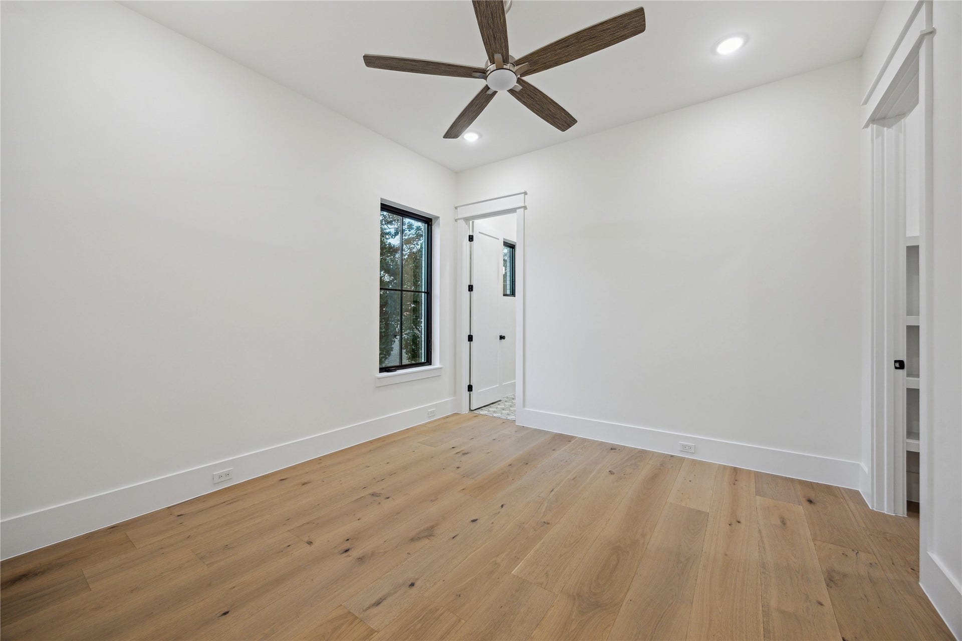 431 W 30th Street, Houston, TX 77018 This room features a minimalist design with white walls, light wood flooring, and a ceiling fan. A large window allows for natural light, and a doorway leads to additional space.