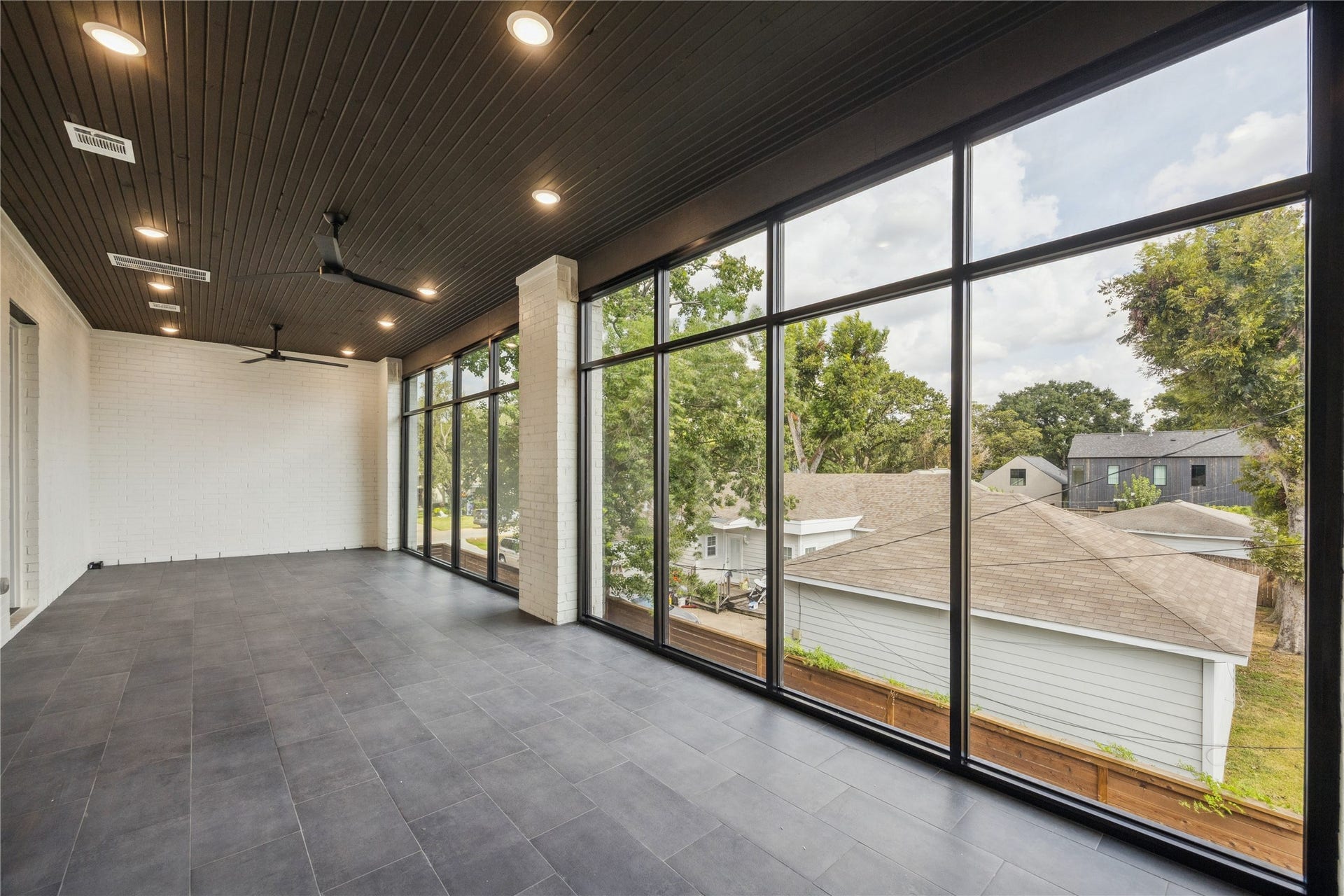 431 W 30th Street, Houston, TX 77018 This photo showcases a spacious sunroom with floor-to-ceiling windows, offering abundant natural light and views of a suburban neighborhood. The room features a modern, dark-tiled floor and a sleek wood-paneled ceiling with recessed lighting and ceiling fans, perfect for relaxation or entertaining.