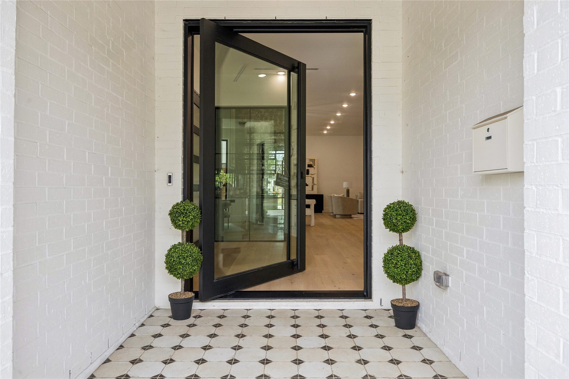 431 W 30th Street, Houston, TX 77018 This photo showcases a modern entryway with a black-framed glass door, flanked by potted topiary plants. The space features a tiled floor with geometric patterns, leading into a bright interior with light wood flooring and contemporary lighting.