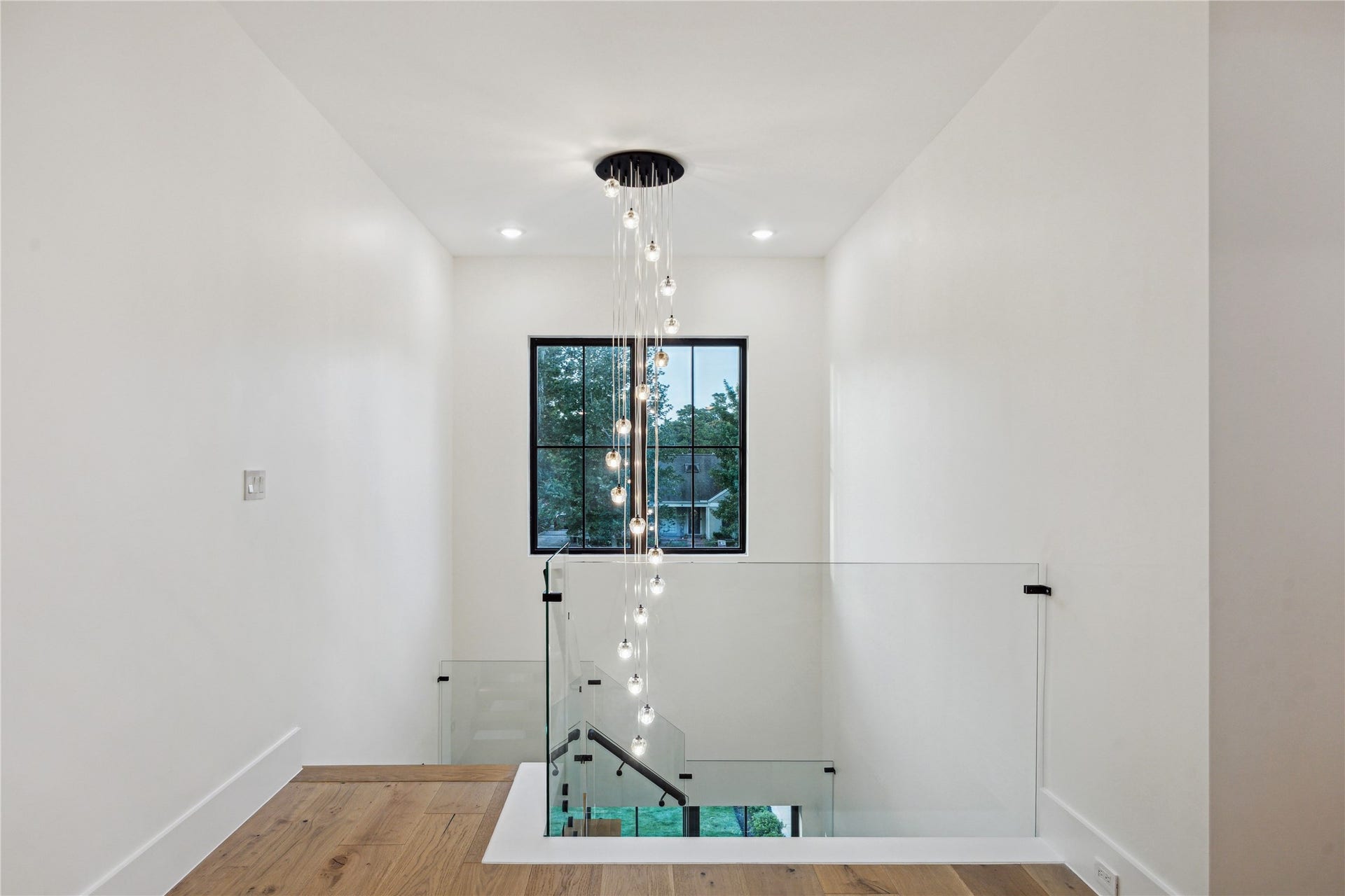 431 W 30th Street, Houston, TX 77018 This photo showcases a modern, bright hallway with a stylish glass railing and a striking pendant light fixture. The space features clean white walls, warm wooden flooring, and a large window offering a view of greenery, creating an inviting and open atmosphere.