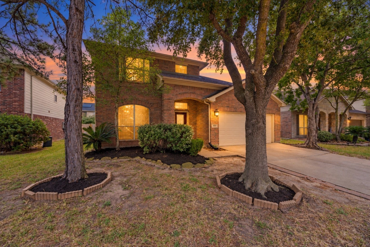 A closer look at the mature front trees and brick facade, with a covered entry set beside the garage and planting beds along the home.