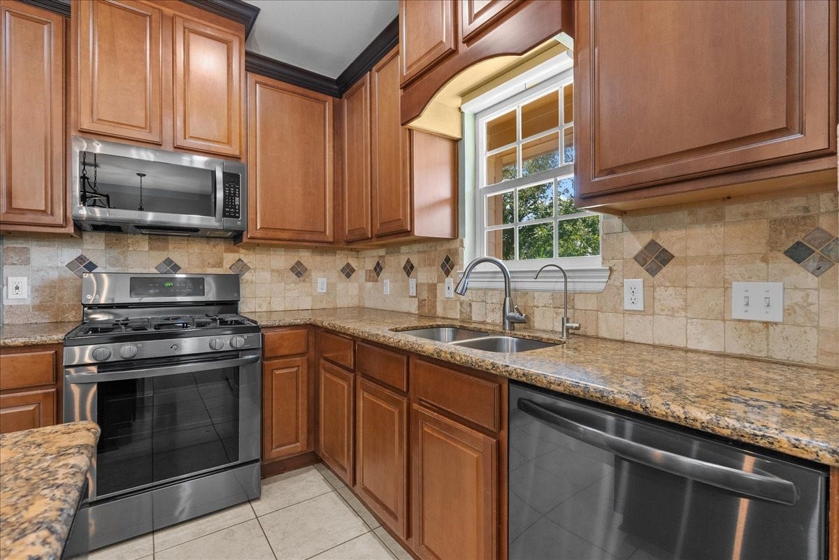 15531 Maple Street, Santa Fe, TX 77517 An arched window frame above the sink brings in natural light and backyard views, paired with a gooseneck faucet and side sprayer. Granite counters wrap the L-shaped layout, with a stainless dishwasher and additional cabinet and drawer storage rounding out the prep space.