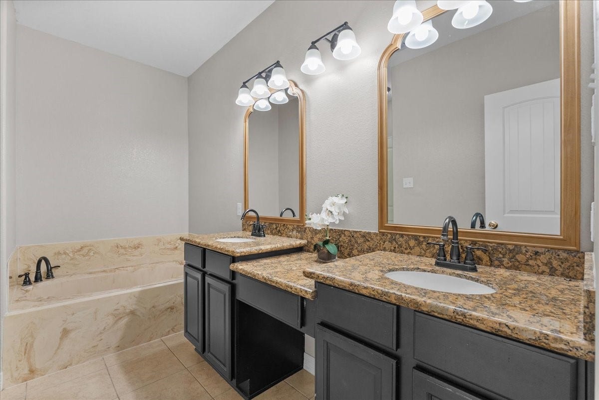 15531 Maple Street, Santa Fe, TX 77517 The primary bath features a double vanity with granite counters and two framed mirrors under wall-mounted light fixtures, plus a separate soaking tub for a spa-like retreat. Dark cabinetry and tile flooring round out the finishes.