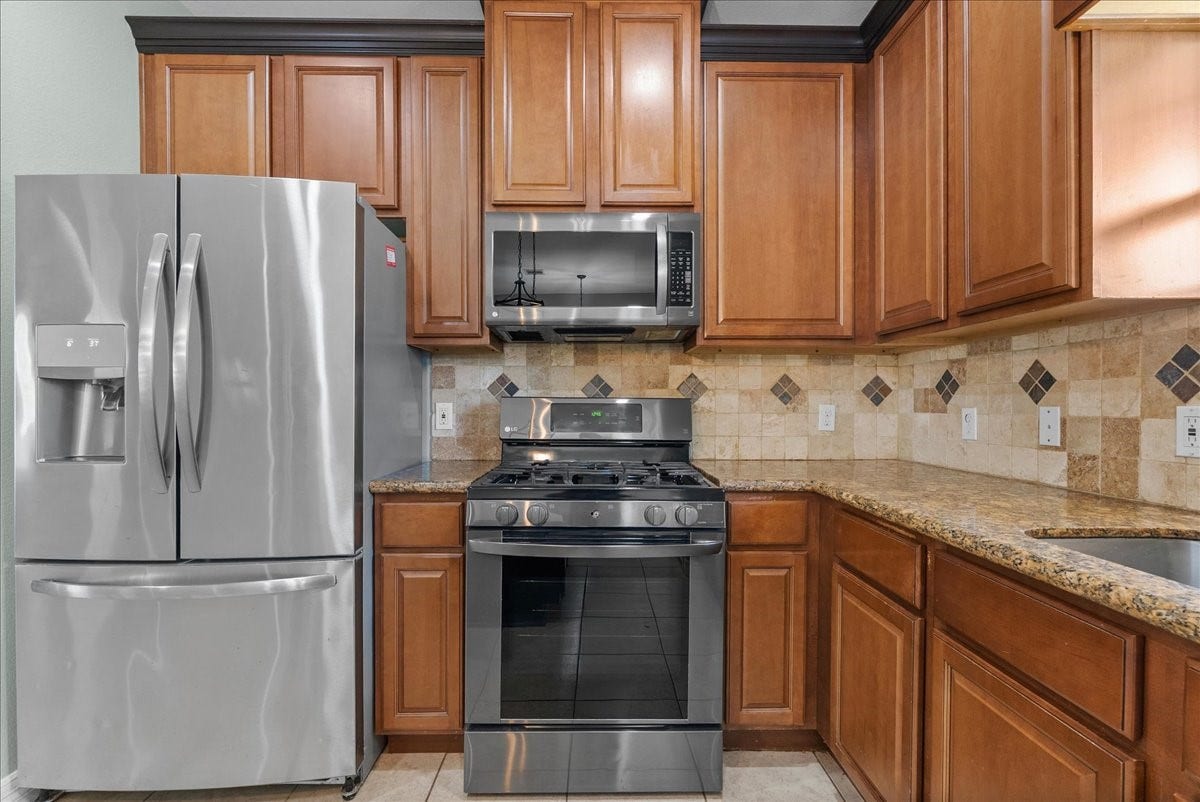 15531 Maple Street, Santa Fe, TX 77517 Stainless steel appliances, a French-door refrigerator with water/ice dispenser, over-the-range microwave, and LG gas range, sit against rich wood cabinetry and granite countertops. A diamond-patterned tile backsplash and matching floor tile add a polished, cohesive finish to the space.