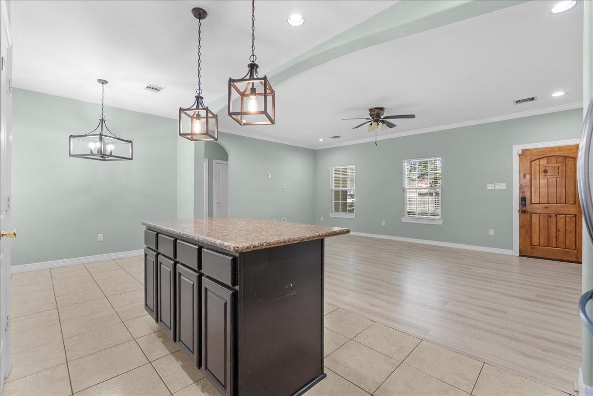 15531 Maple Street, Santa Fe, TX 77517 Three pendant fixtures illuminate the granite island, which opens directly to the vaulted living area beyond. Wide-plank flooring picks up where the kitchen tile leaves off, paired windows bring in natural light, and a ceiling fan plus the wood front entry door anchor the far end of the room.