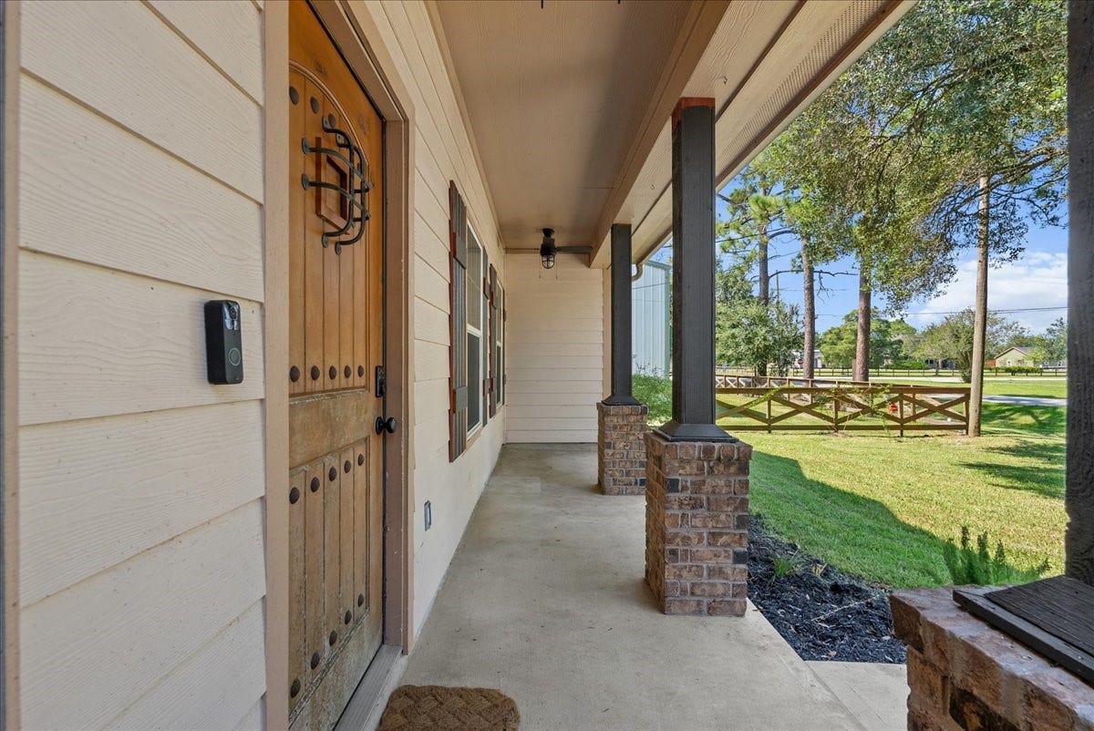 15531 Maple Street, Santa Fe, TX 77517 The arched wood door with wrought-iron hardware and a video doorbell sets a secure, custom tone right at the threshold, while the long covered walkway connects seamlessly to the porch beyond, framing clean sightlines out to the fenced yard and treeline.