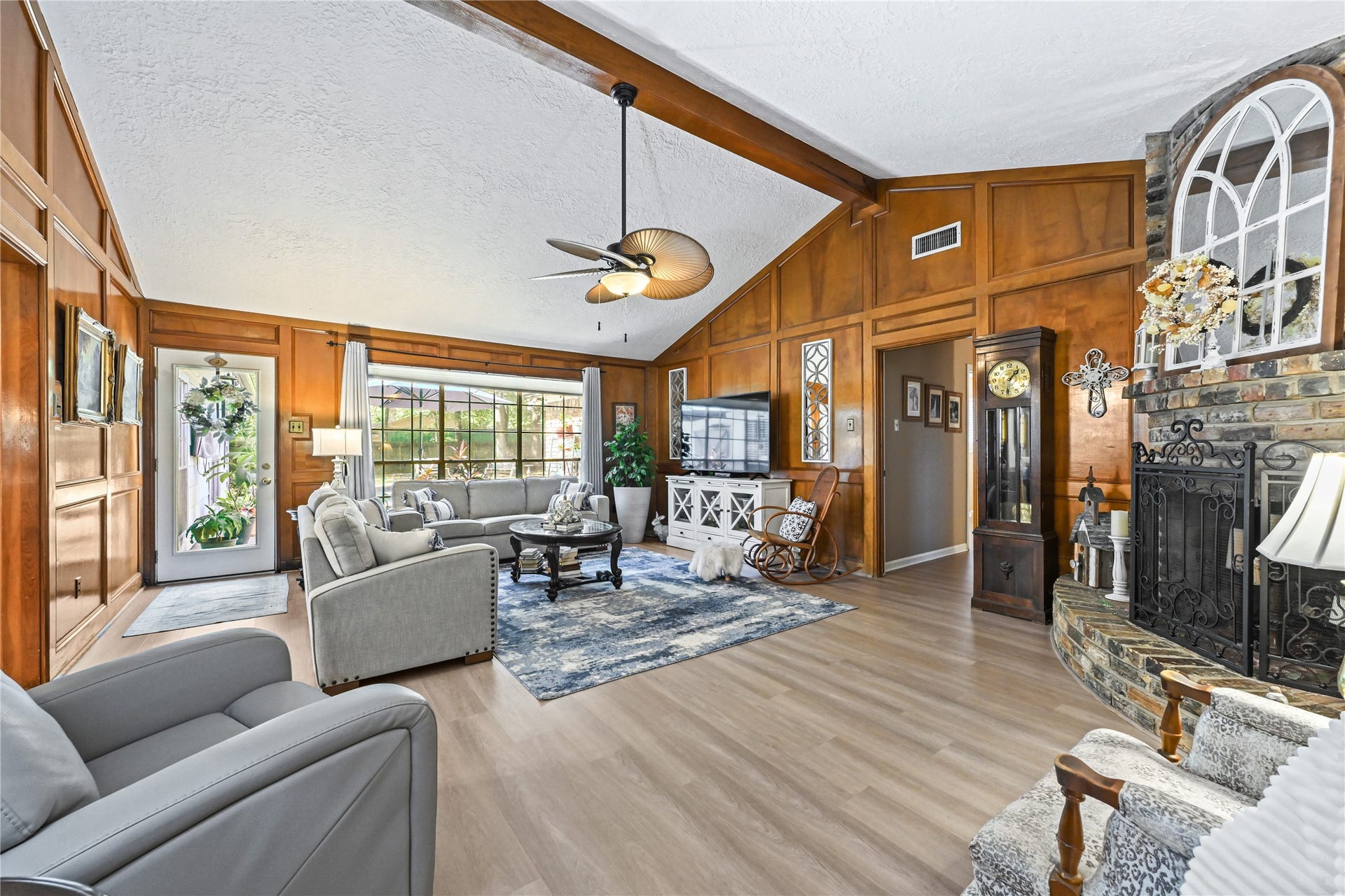 25803 Maplewood Drive, Spring, TX 77386 Spacious living room with vaulted ceilings, rich wood paneling, and a cozy brick fireplace. Features large windows for natural light, elegant furnishings, and a stylish ceiling fan, creating a warm and inviting atmosphere.