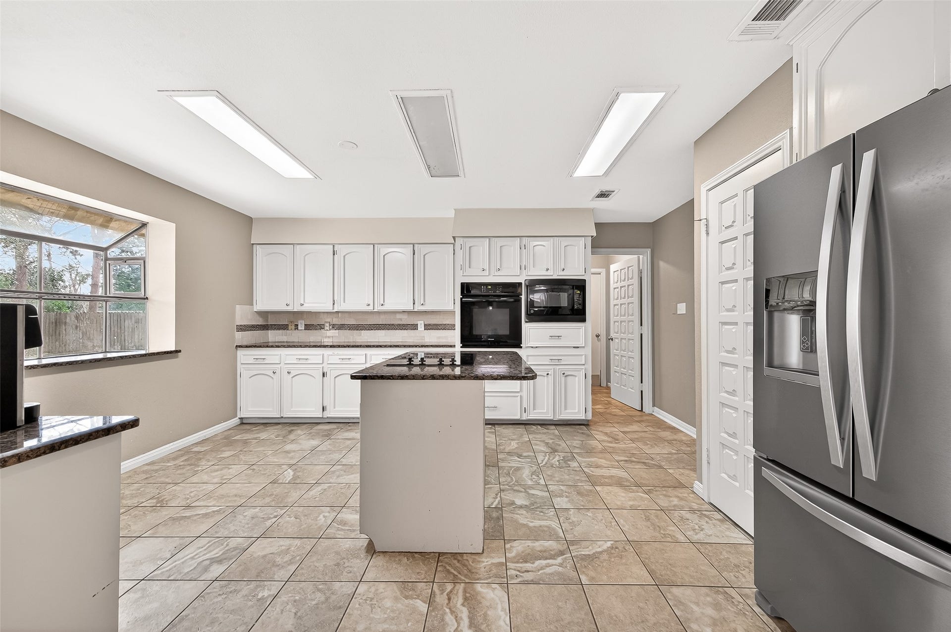 5607 Avenue M 1/2, Santa Fe, TX 77510 This large kitchen features modern appliances, ample white cabinetry, and a central island with a cooktop. Natural light flows through a large window, highlighting the tiled floor and a contemporary design perfect for cooking and entertaining.