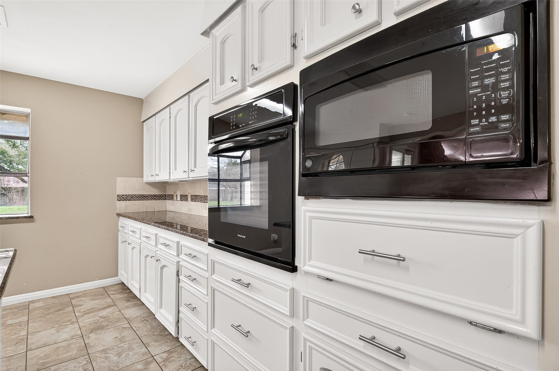 5607 Avenue M 1/2, Santa Fe, TX 77510 This kitchen features modern black appliances, including a built-in oven and microwave, set against crisp white cabinetry. The tile countertops and backsplash add a touch of elegance, while the large window provides ample natural light.