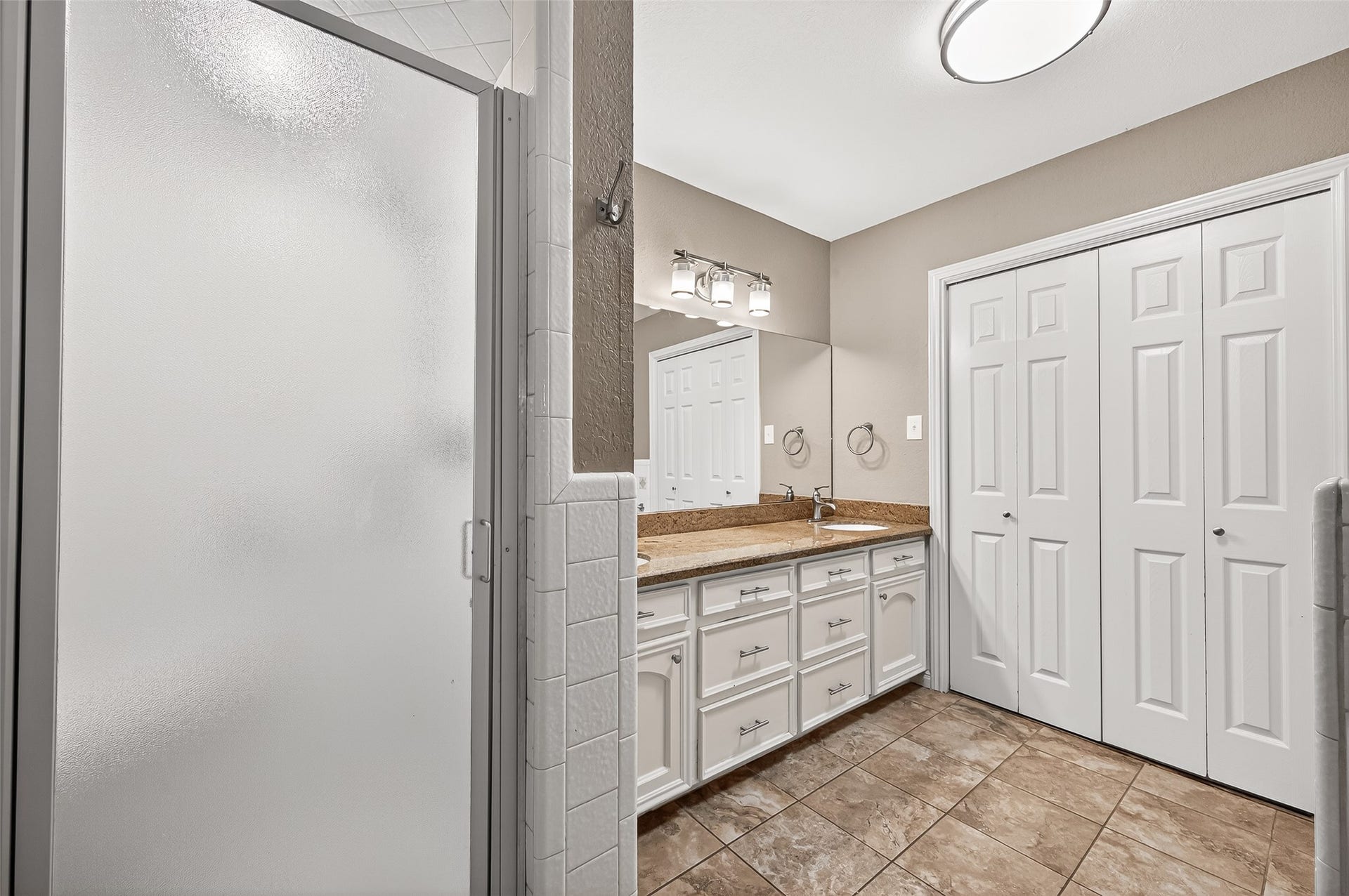 5607 Avenue M 1/2, Santa Fe, TX 77510 This bathroom features a modern frosted glass shower, double vanity with granite countertops, ample cabinet storage, and large closet doors, complemented by tile flooring and sleek lighting.