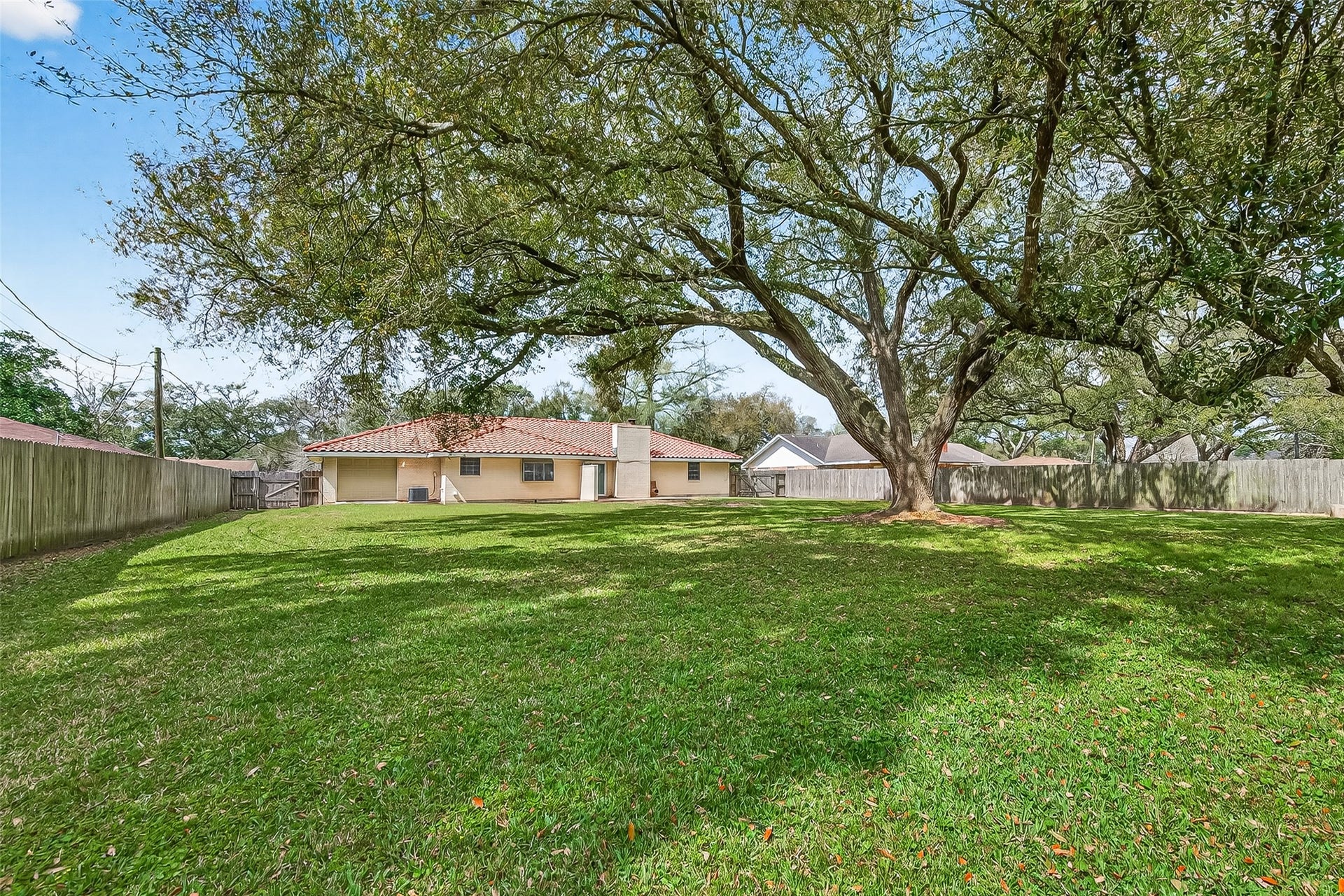 5607 Avenue M 1/2, Santa Fe, TX 77510 This photo showcases a spacious backyard with a large, mature tree providing ample shade. The area is fully fenced, offering privacy, and the view includes a single-story house with a red-tiled roof, making it ideal for outdoor activities and relaxation.