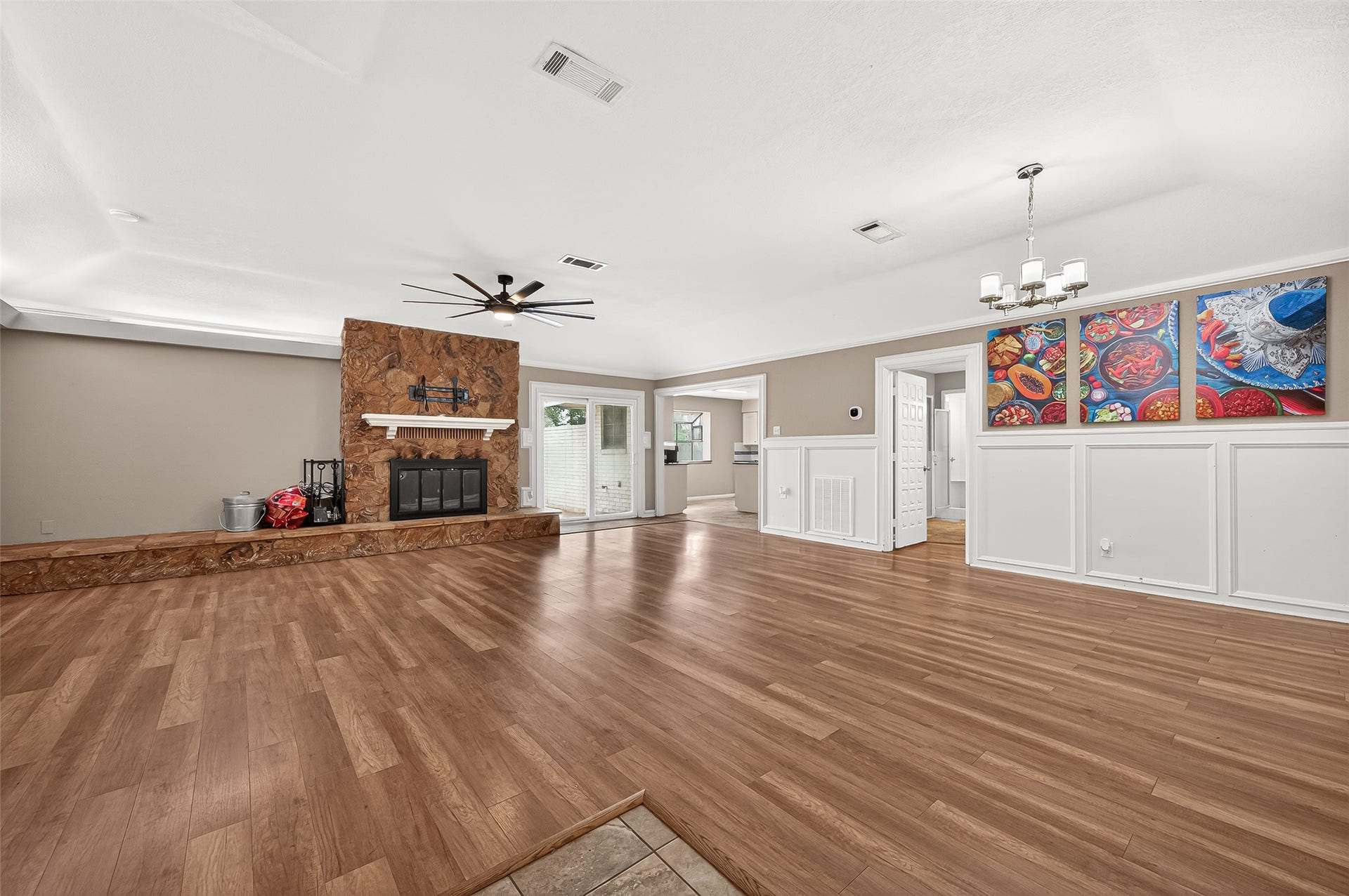 5607 Avenue M 1/2, Santa Fe, TX 77510 This spacious living area features hardwood flooring and a stone fireplace. It includes a ceiling fan, dining space with a modern chandelier, and vibrant wall art. The room opens to a bright kitchen, offering a welcoming, open-concept feel.