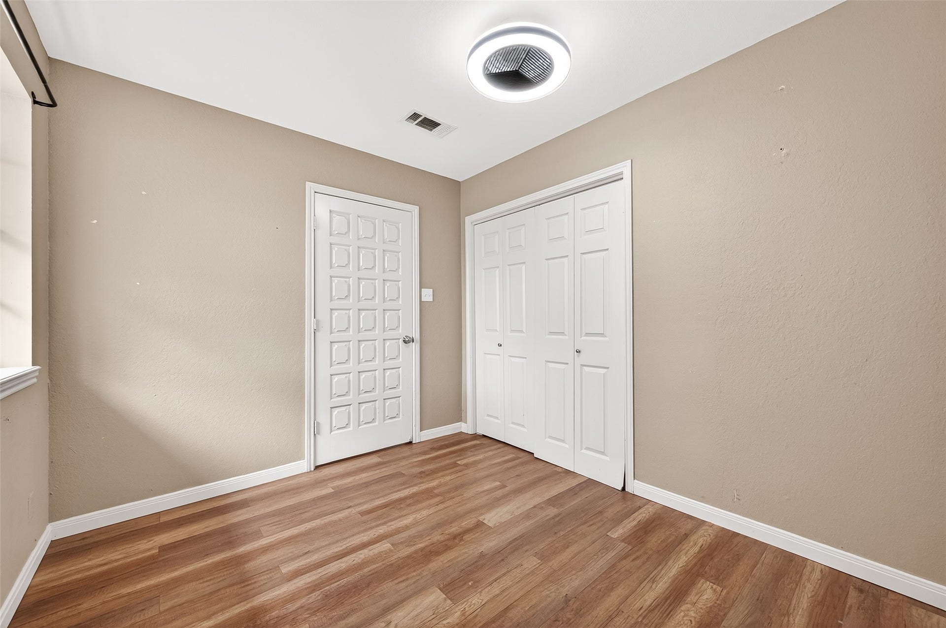 5607 Avenue M 1/2, Santa Fe, TX 77510 This room features light brown walls and laminate wood flooring, with a window allowing natural light. It includes a modern ceiling light and two white doors, one with a geometric design and the other a closet, providing ample storage space.