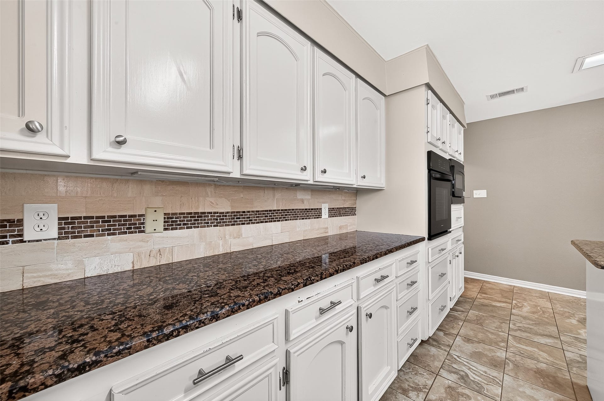 5607 Avenue M 1/2, Santa Fe, TX 77510 This kitchen features elegant white cabinetry with ample storage, stunning dark granite countertops, and a stylish tile backsplash. The space is complemented by tiled flooring and built-in appliances, offering a modern and functional layout.