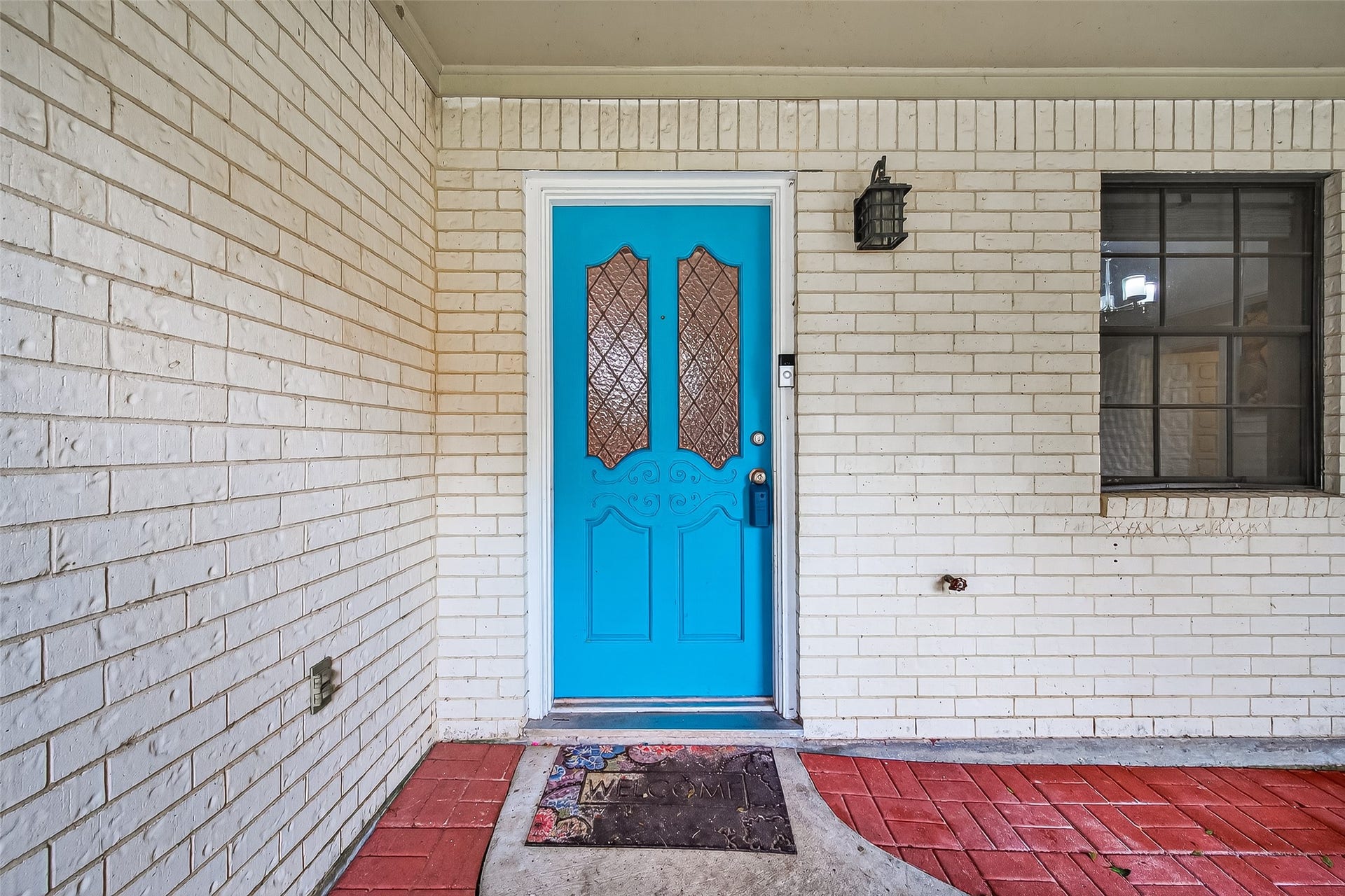 5607 Avenue M 1/2, Santa Fe, TX 77510 This photo showcases a welcoming front entrance with a vibrant blue door set in a white brick wall. The entrance features a decorative glass panel, a black outdoor light fixture, and a red brick pathway leading to a "Welcome" mat. A window with black trim is visible beside the door.
