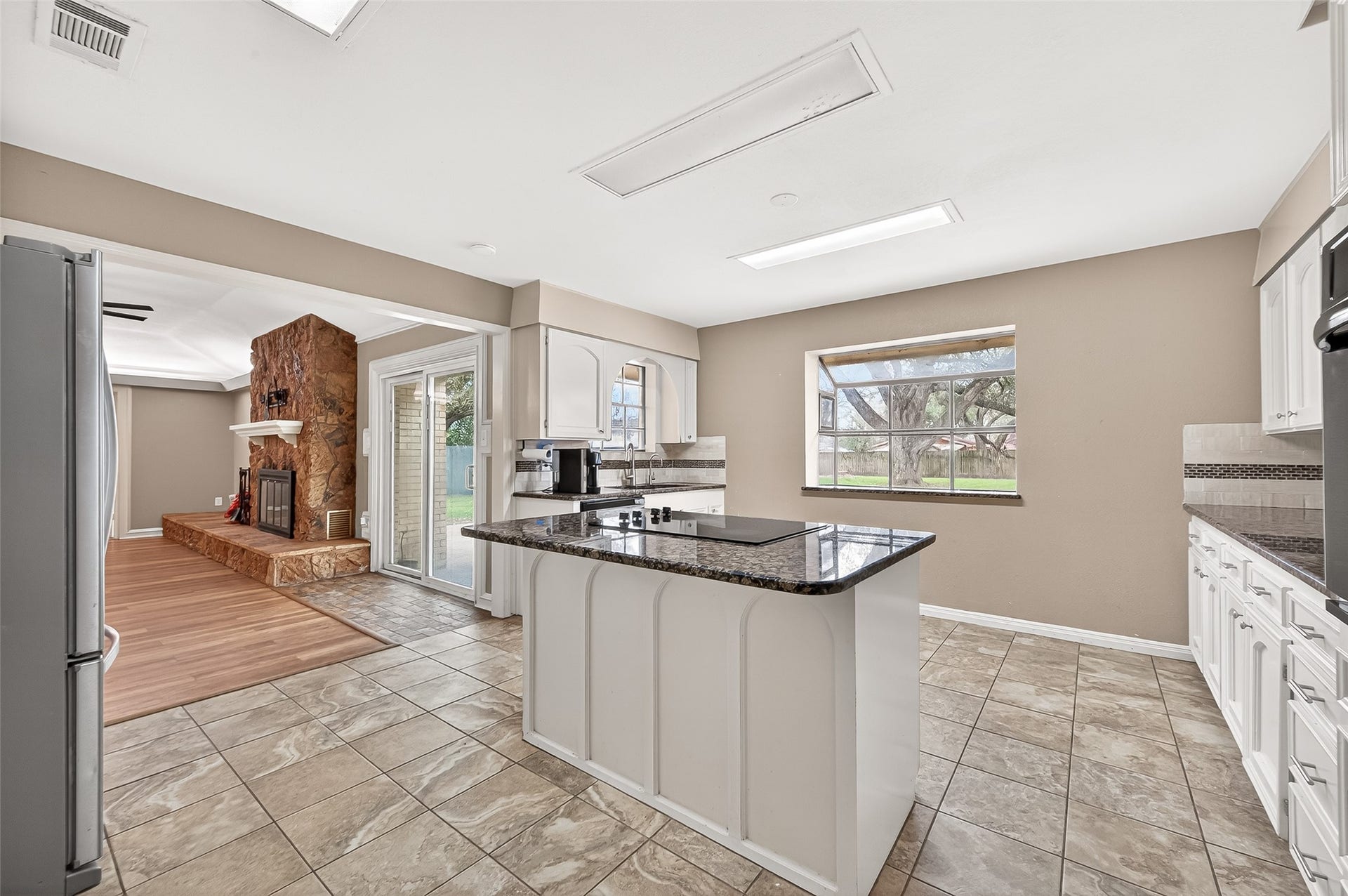 5607 Avenue M 1/2, Santa Fe, TX 77510 This kitchen features a central island with a cooktop, granite countertops, and ample cabinetry. It opens to a cozy living area with a stone fireplace and sliding doors leading to the backyard, offering natural light and a seamless flow for entertaining.