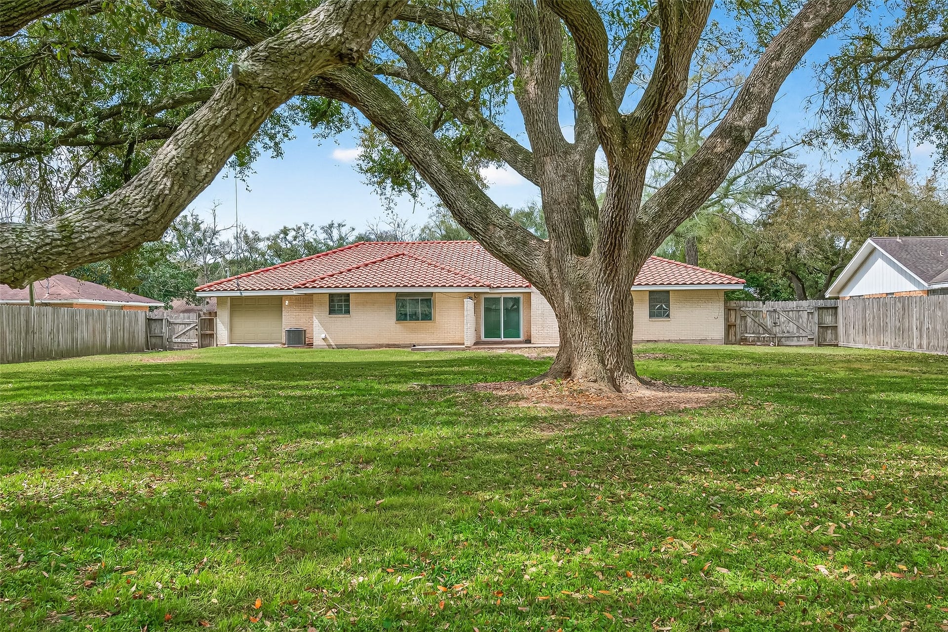 5607 Avenue M 1/2, Santa Fe, TX 77510 This photo features a charming single-story home with a red-tiled roof, set in a spacious, well-maintained backyard. A large, mature tree provides shade, and the yard is enclosed by a wooden fence, offering privacy and ample space for outdoor activities.