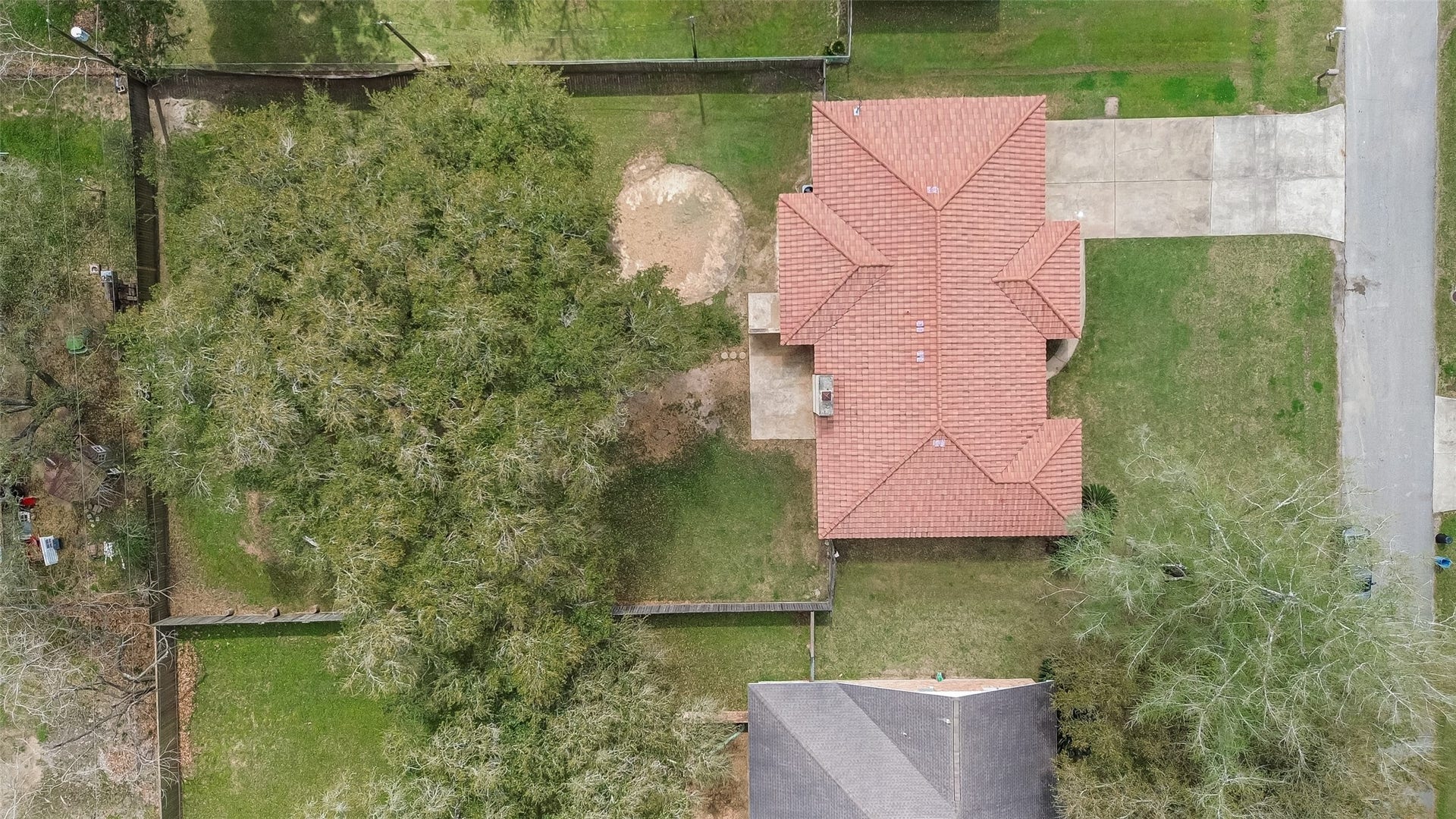 5607 Avenue M 1/2, Santa Fe, TX 77510 This aerial view shows a single-story home with a red-tiled roof and a spacious driveway. The property features a large fenced backyard with mature trees, providing ample shade and privacy. The house is situated in a residential area with nearby green spaces.