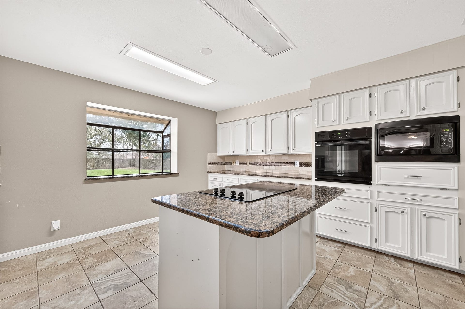 5607 Avenue M 1/2, Santa Fe, TX 77510 This spacious kitchen features a large window providing ample natural light, granite countertops, white cabinetry, and modern appliances, including a built-in oven and microwave. The central island offers additional prep space and a cooktop, perfect for culinary enthusiasts.