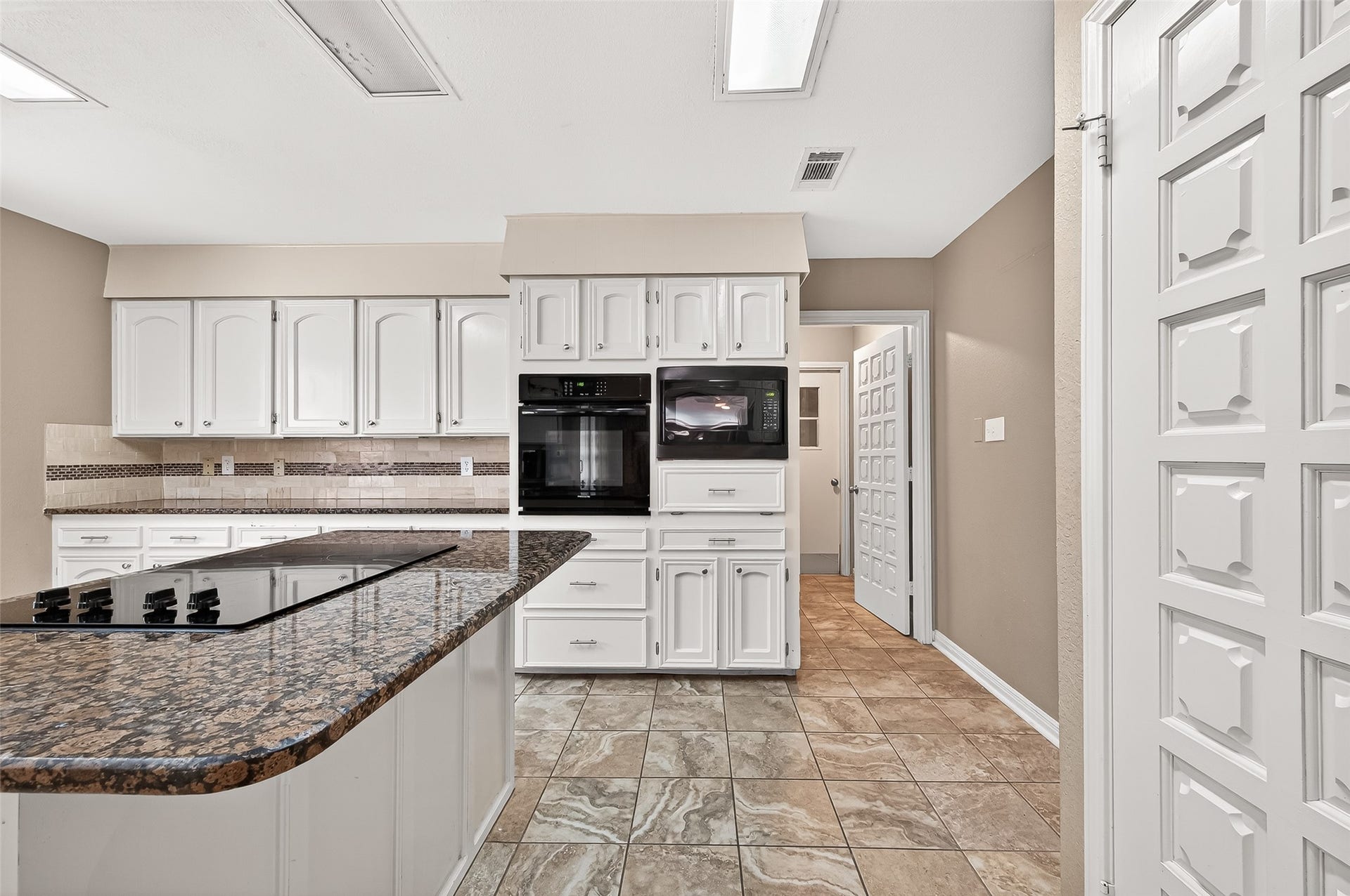 5607 Avenue M 1/2, Santa Fe, TX 77510 This kitchen features white cabinetry, a built-in oven and microwave, and a granite countertop island with a cooktop. The neutral tile flooring and backsplash add a classic touch, while the paneled doors provide a cohesive look.