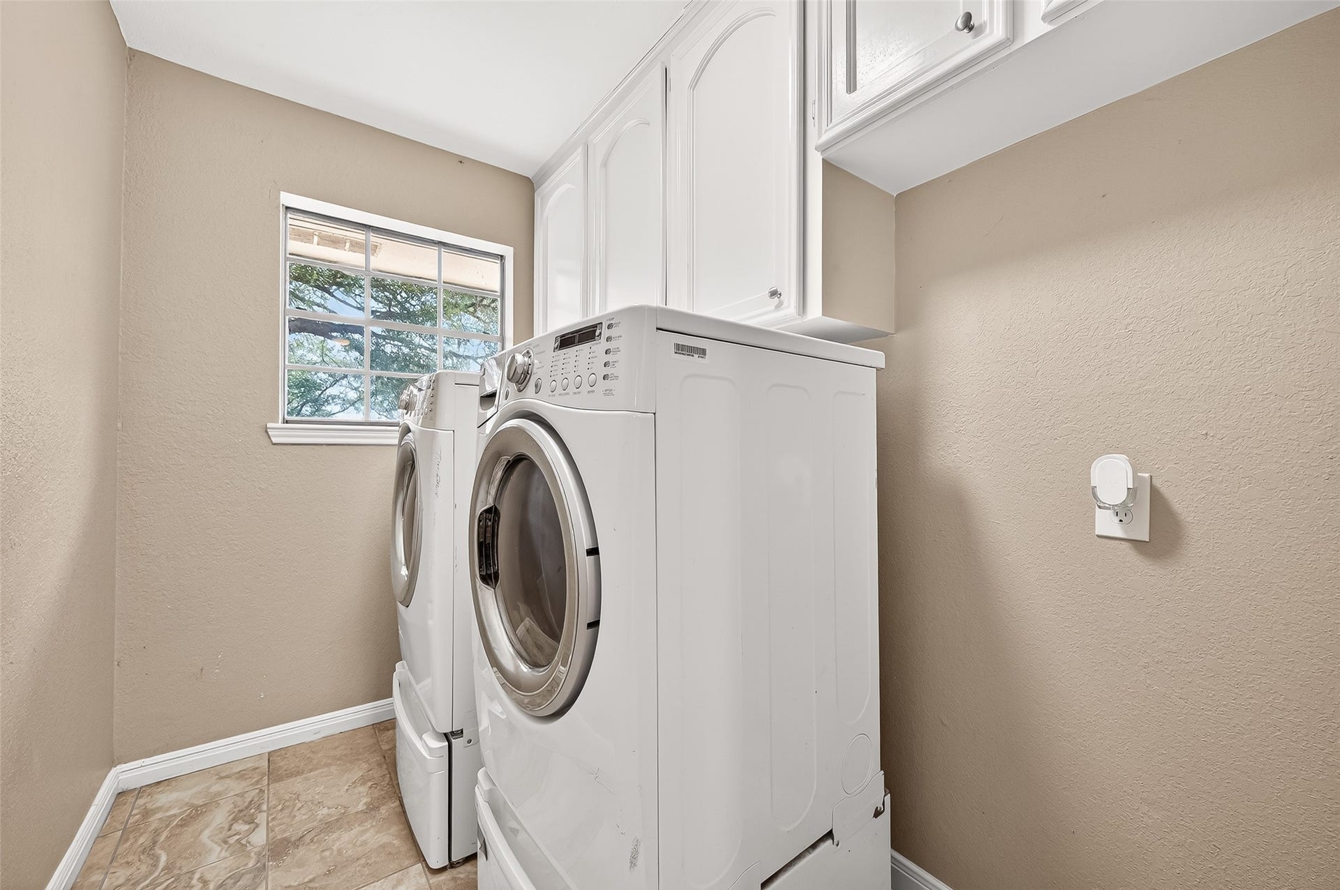 5607 Avenue M 1/2, Santa Fe, TX 77510 This photo shows a compact laundry room with a front-loading washer and dryer (washer and dryer not included). It features beige walls, white cabinetry for storage, and a window providing natural light. The floor is tiled, offering easy maintenance.