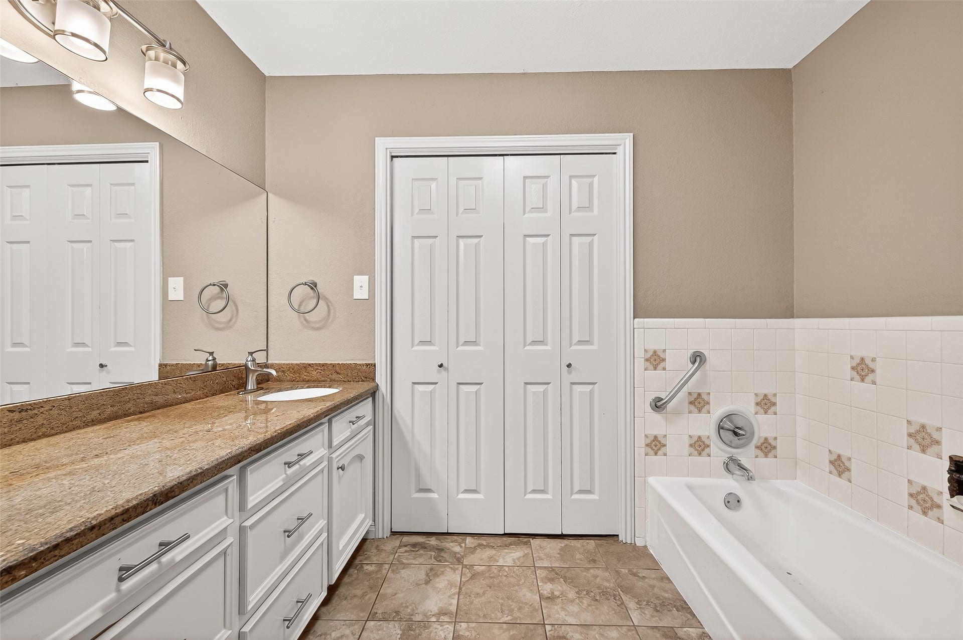 5607 Avenue M 1/2, Santa Fe, TX 77510 This bathroom features a long granite countertop with a single sink, ample storage with white cabinets, a tiled bathtub with a grab bar, and a double door closet. The decor is neutral, providing a clean and spacious feel.